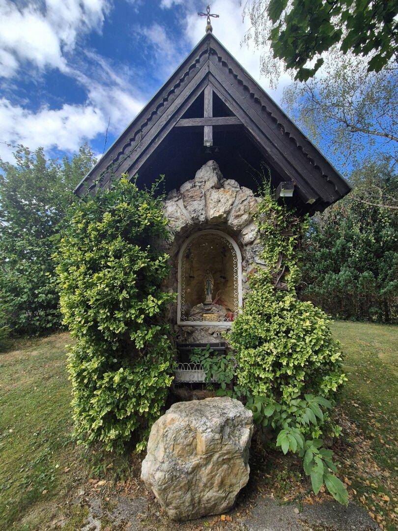 Kleine, traditionelle Kapelle mit Steindekoration und grünen Büschen in ländlicher Umgebung.