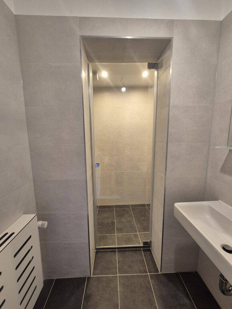 Modernes Badezimmer mit grauen Fliesen, einer Duschkabine und einem einzelnen Waschbecken.