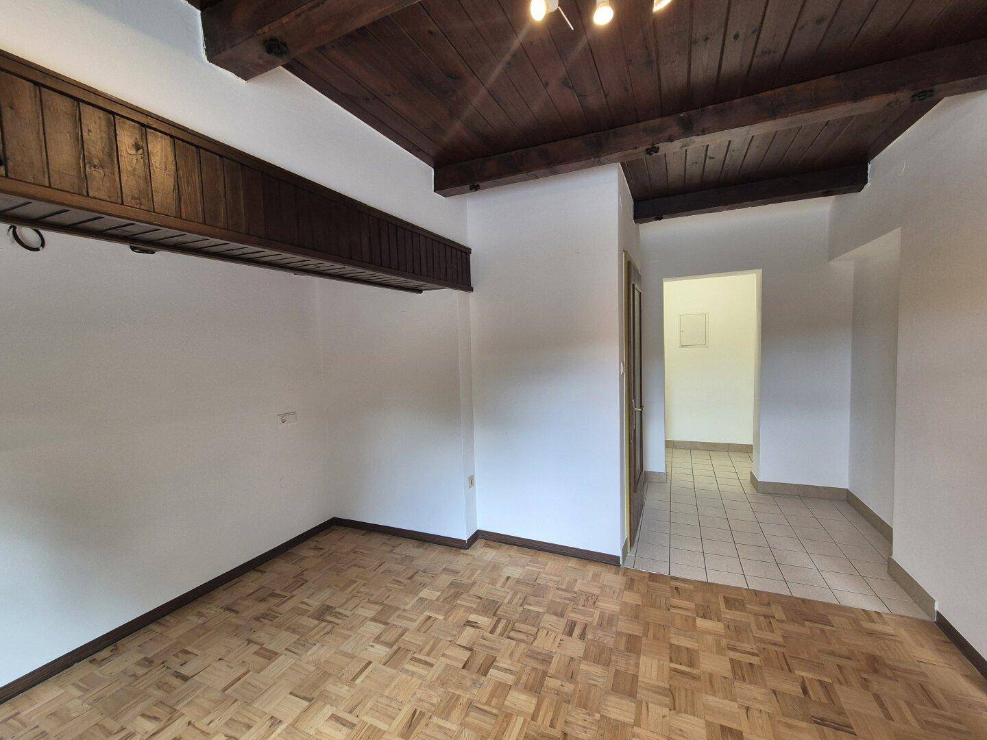 Heller Wohnraum mit Parkettboden und dunklen Holzdeckenbalken, der in einen gefliesten Flur übergeht.