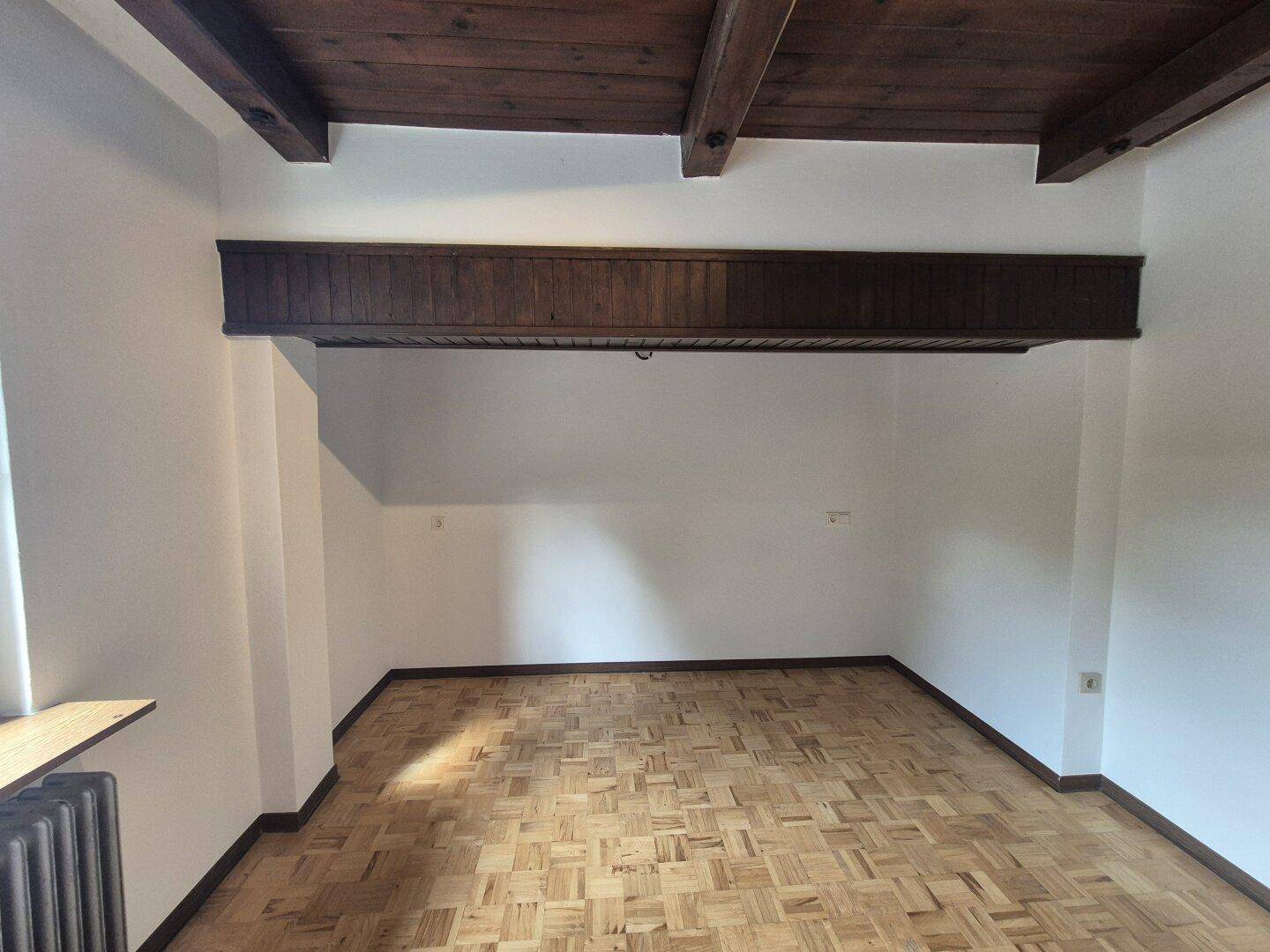 Geräumiger Raum mit Fischgrätparkett, weißen Wänden und einer rustikalen Holzdeckenbalkenkonstruktion.