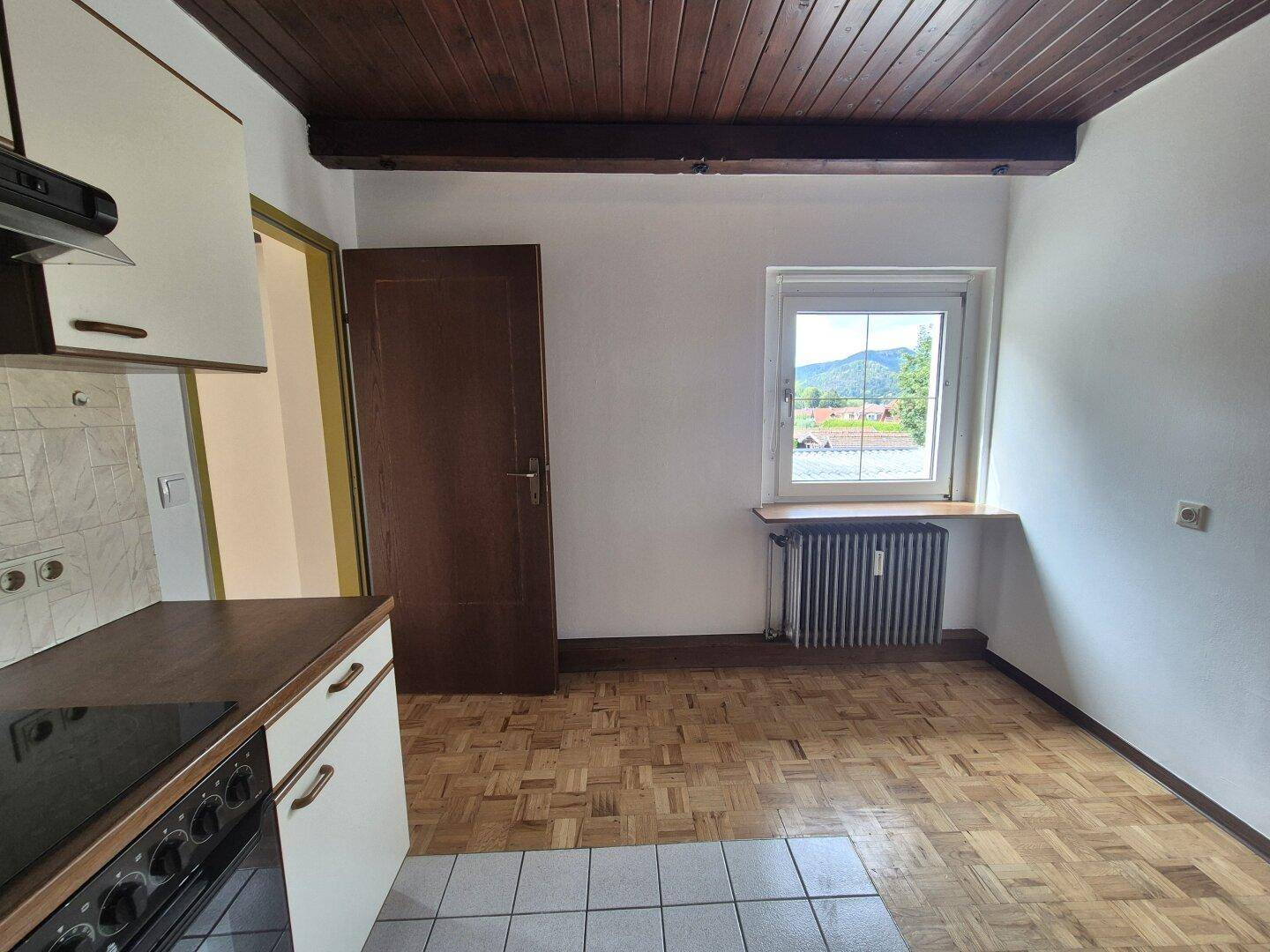 Küchenbereich mit Fenster und Heizkörper, Parkettboden und Blick auf die umliegende Landschaft.