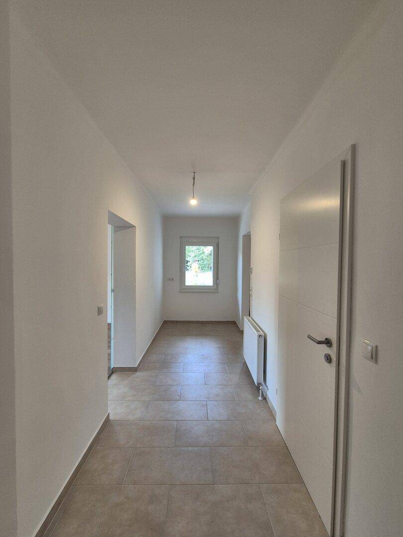 Langer, heller Flur mit Fliesenboden, Fenster und Heizkörper.
