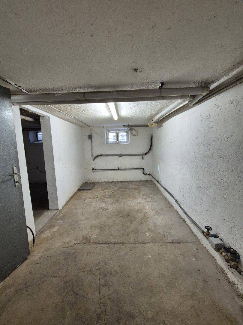 Langer Kellerraum mit Betonboden, Fenster und freiliegenden Leitungen.