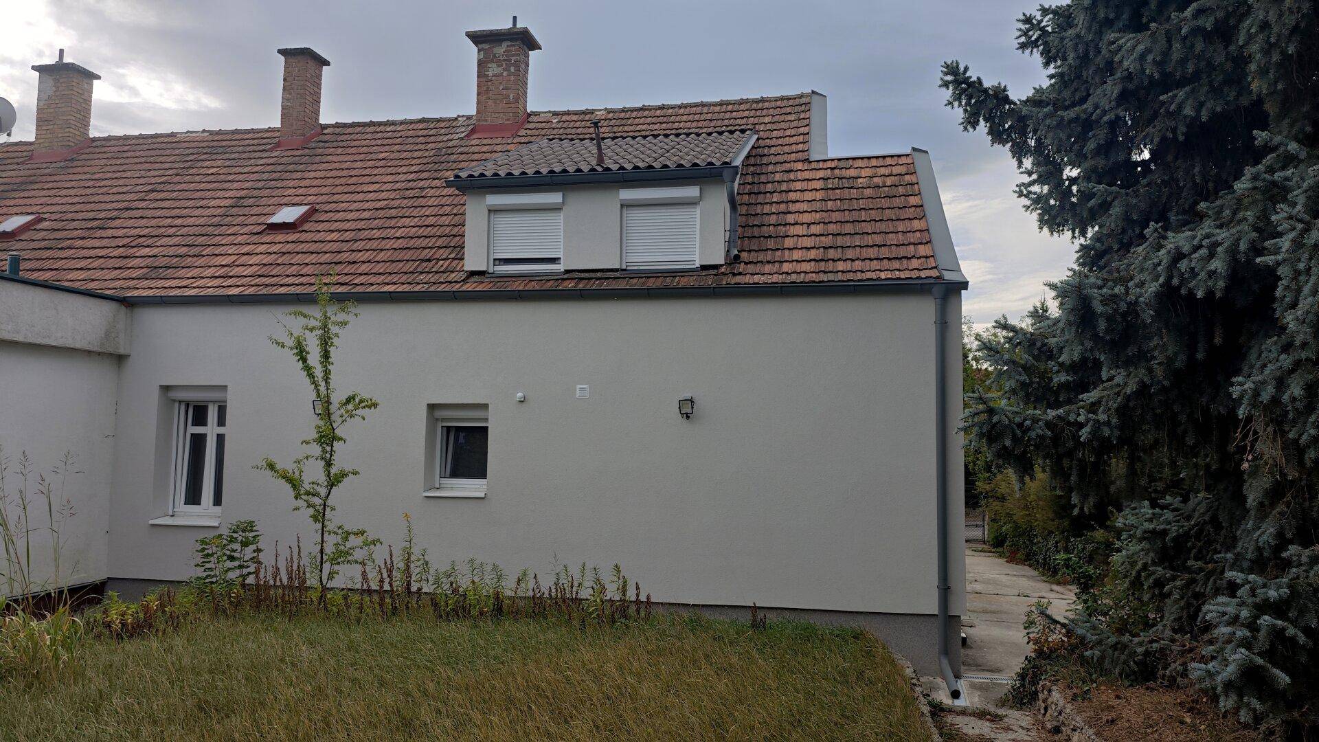 Seitenansicht des Hauses mit Dachgaube, Rollläden und Bäumen im Hintergrund.