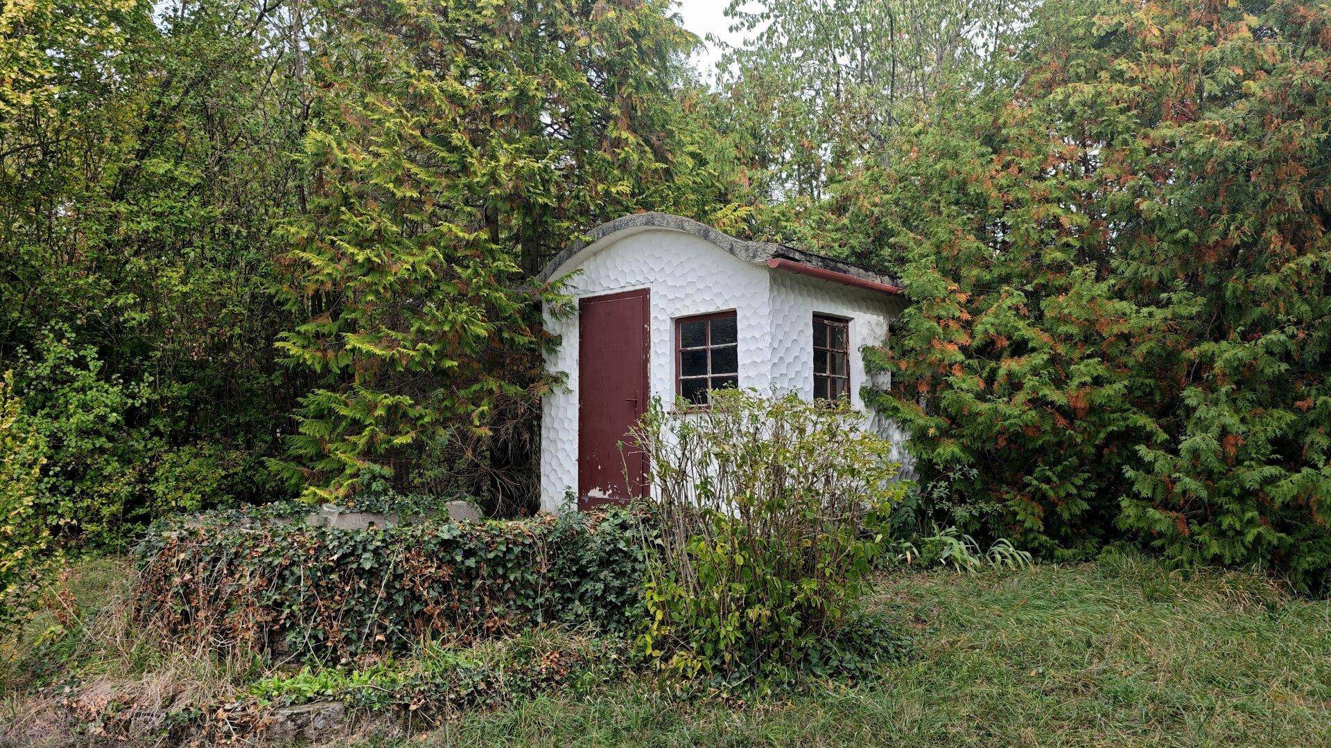 Kleines, weiß verputztes Gartenhaus mit roter Tür und zwei Fenstern, eingebettet in dichte Bäume und Sträucher.