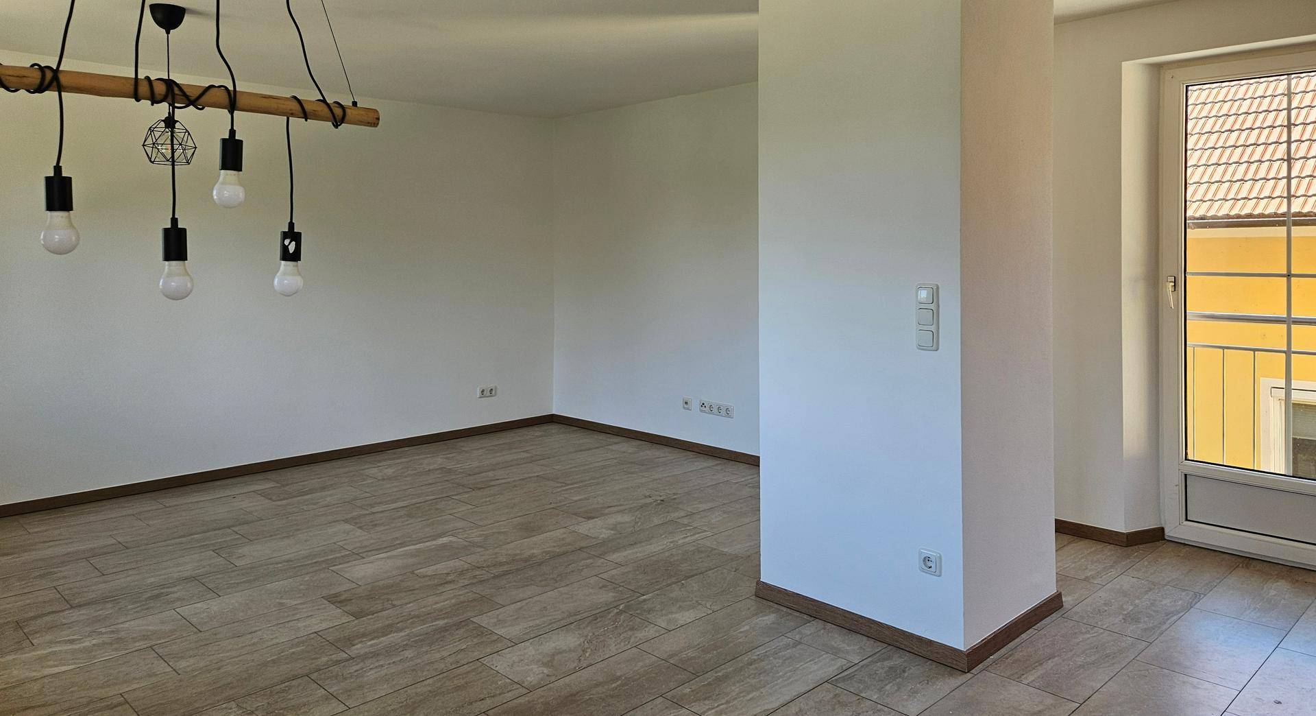 Heller, leerer Wohnbereich mit Fliesenboden und Zugang zu einem Balkon oder einer Terrasse.