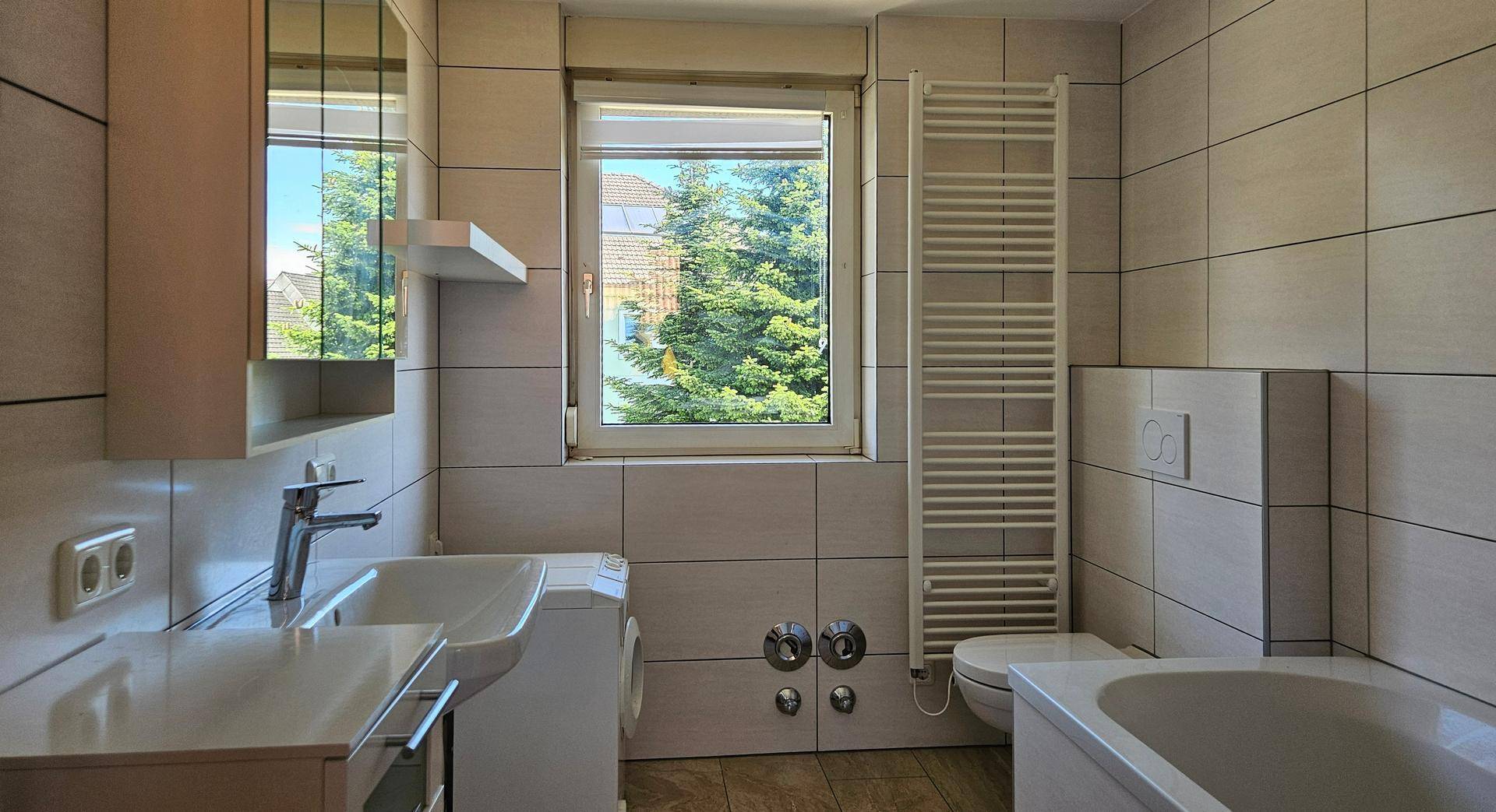 Gefliestes Badezimmer mit Fenster, Waschbecken, Toilette, Badewanne und Handtuchheizkörper.