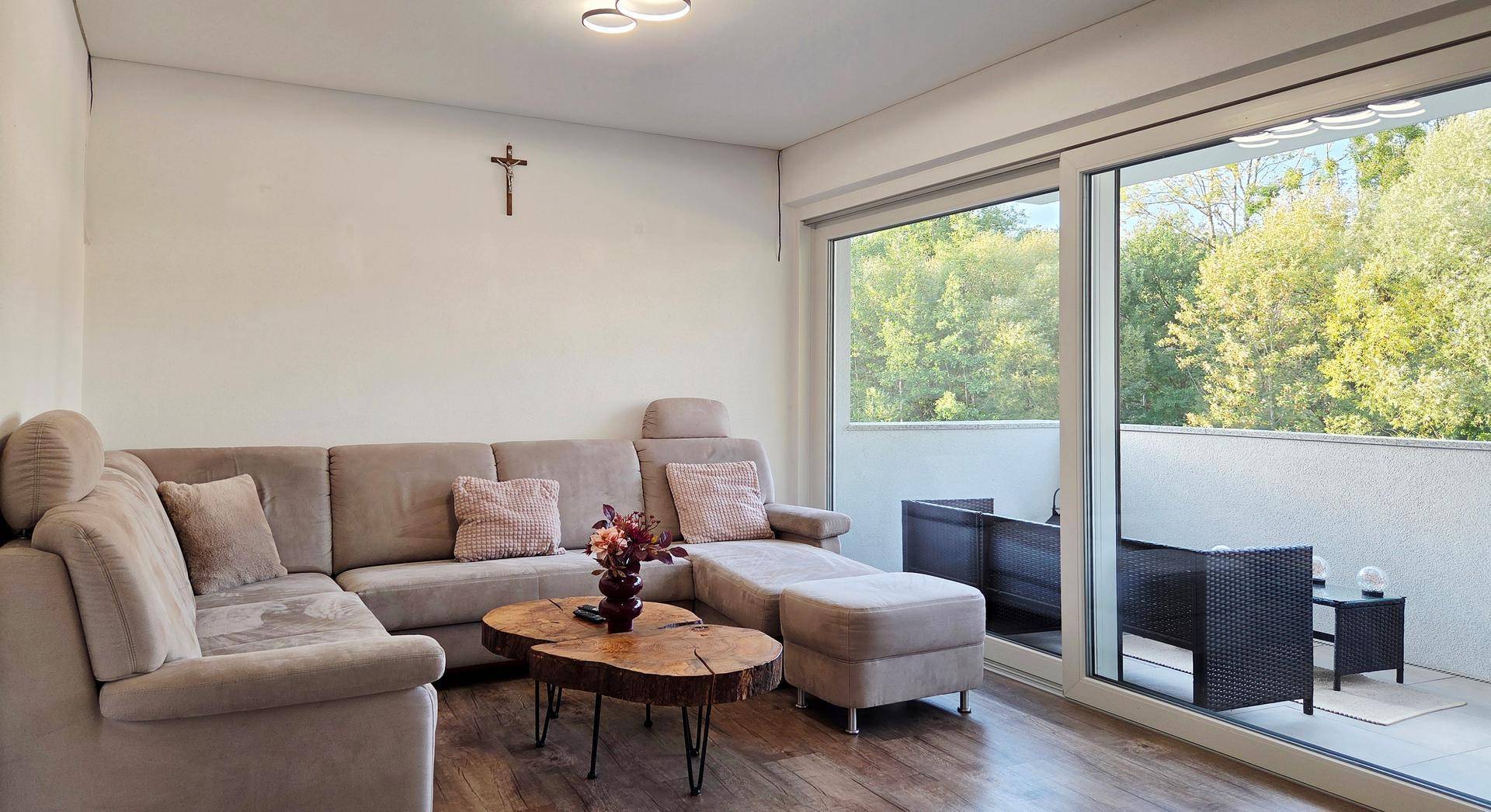 Gemütliches Wohnzimmer mit großem Ecksofa, Holztisch und Zugang zu einem Balkon mit Grünblick.