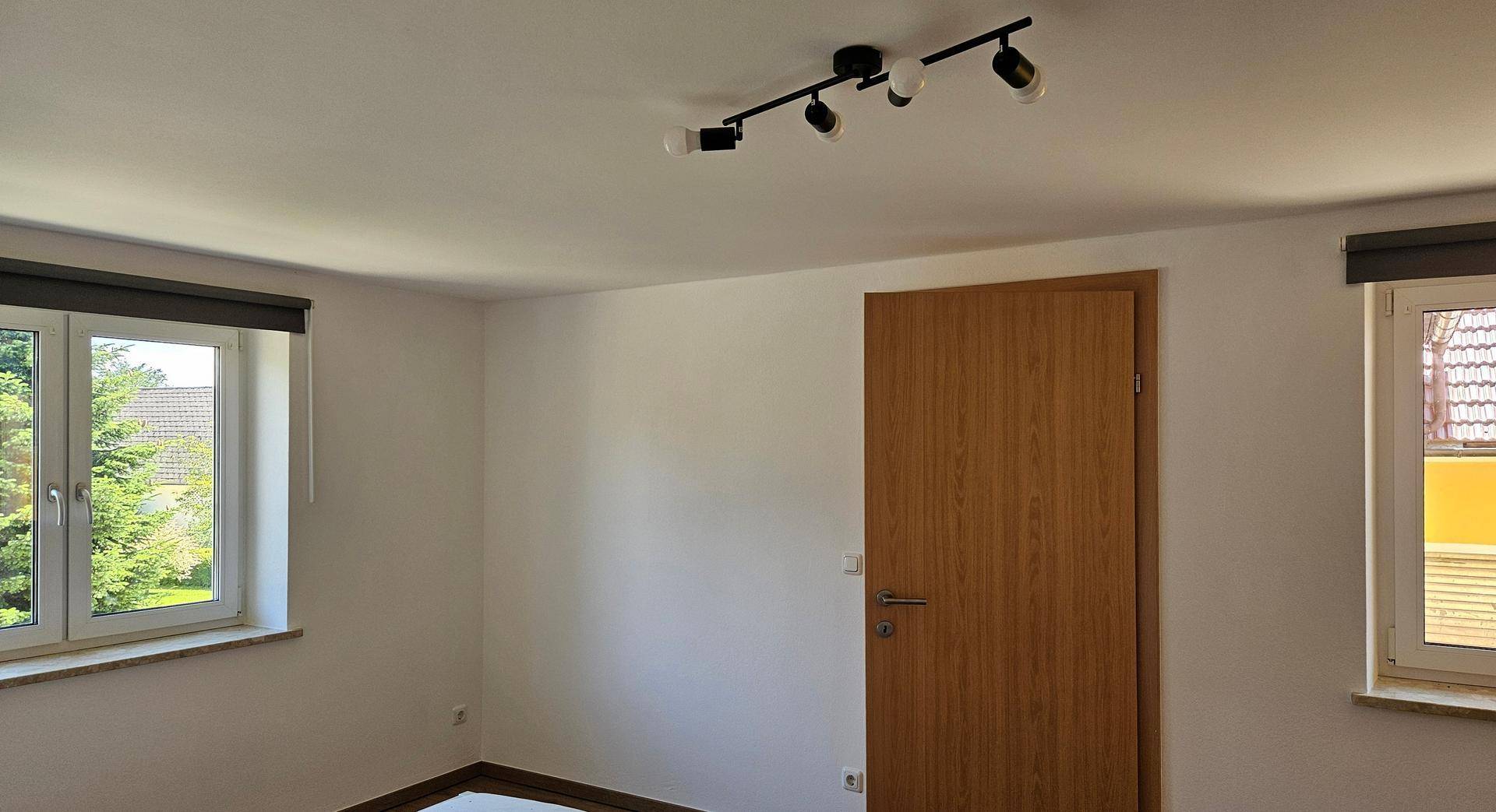 Helles Schlafzimmer mit zwei Fenstern, Holztür und einfacher Deckenbeleuchtung.