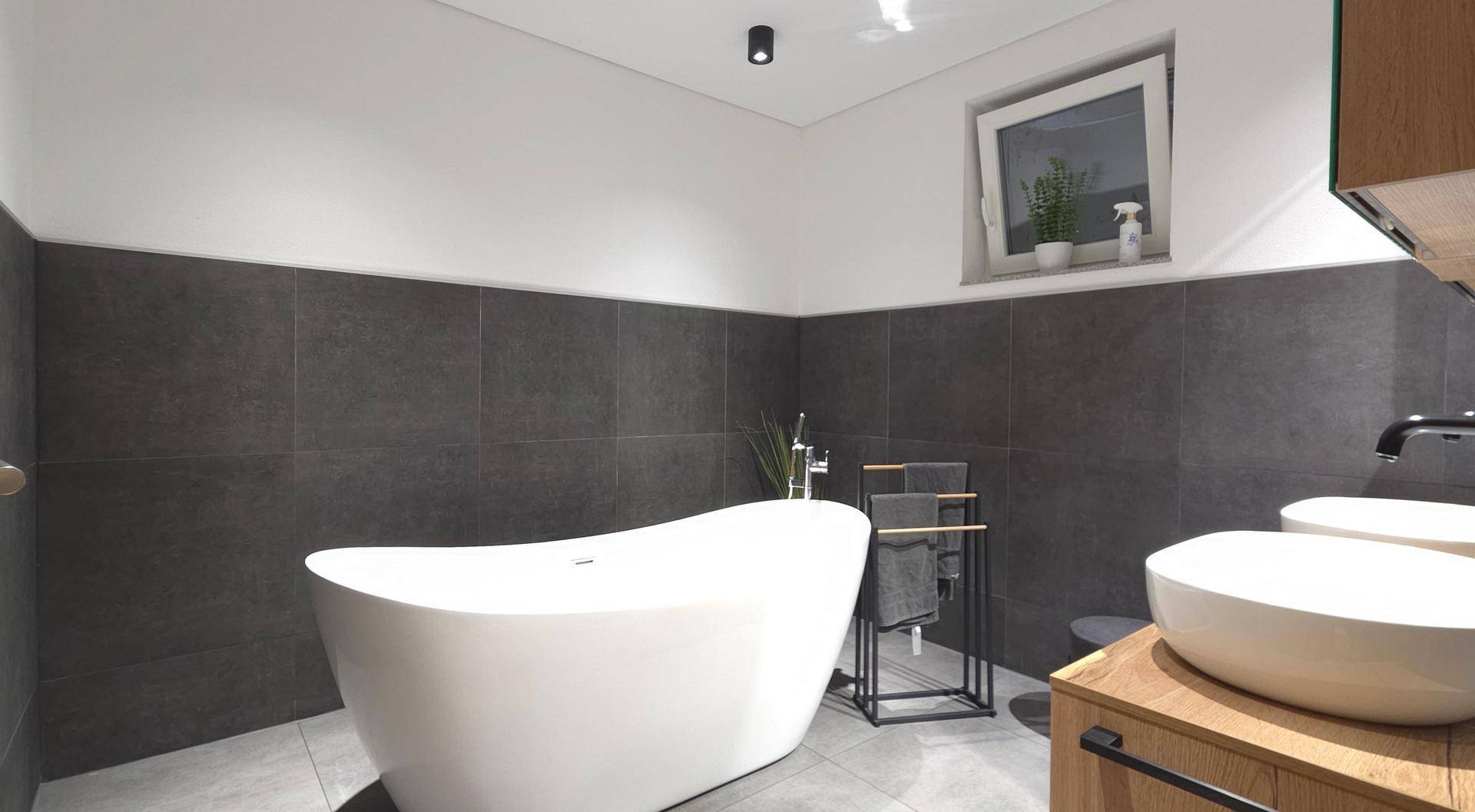 Elegantes Badezimmer mit freistehender Badewanne, dunklen Fliesen und einem Fenster für Tageslicht.