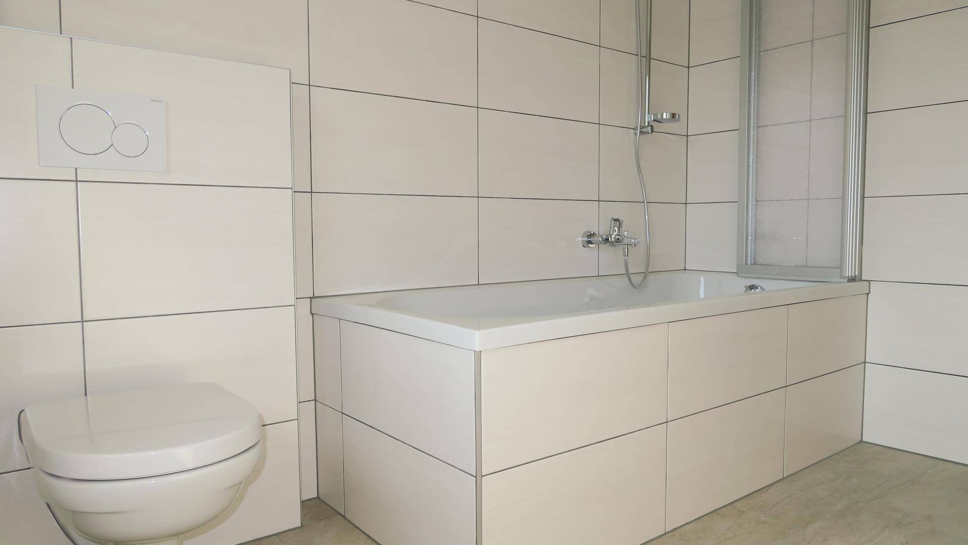 Modern gefliestes Badezimmer mit Badewanne inklusive Duschabtrennung und wandhängendem WC.