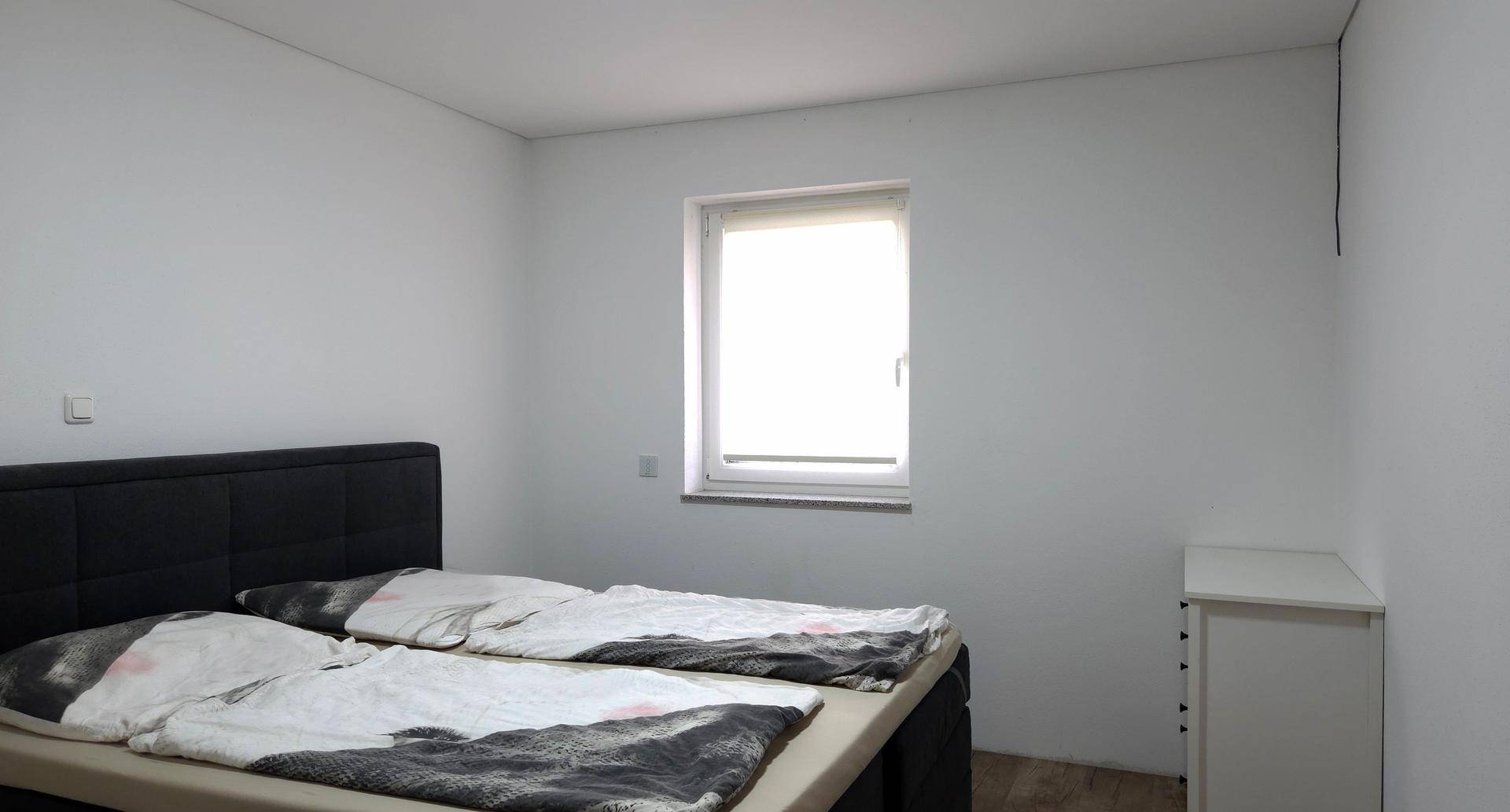 Helles Schlafzimmer mit Doppelbett, weißer Kommode und einem Fenster für natürliches Licht.