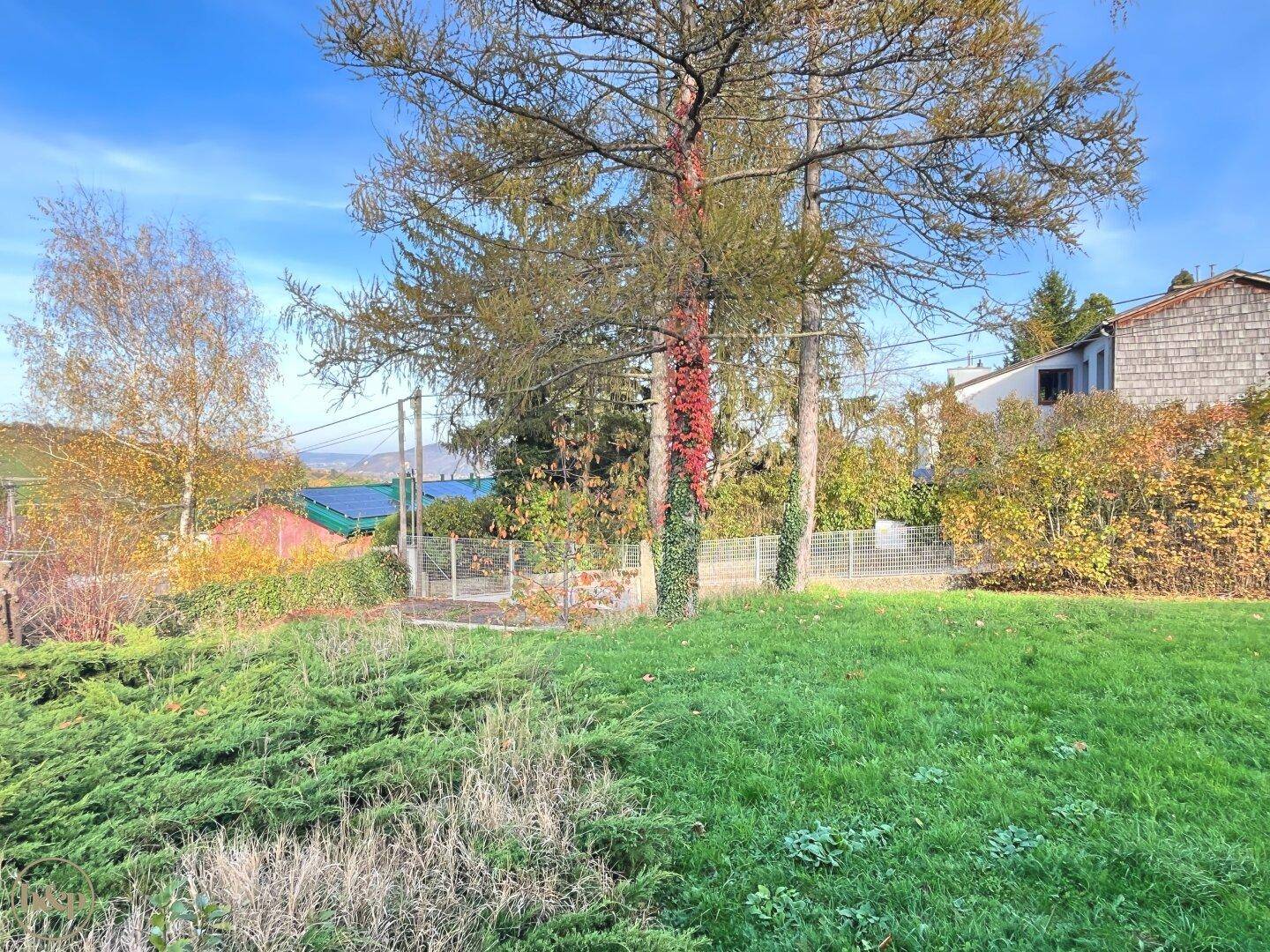 Garten mit Rasenfläche und Hecken, im Hintergrund sind weitere Häuser und die Landschaft sichtbar.
