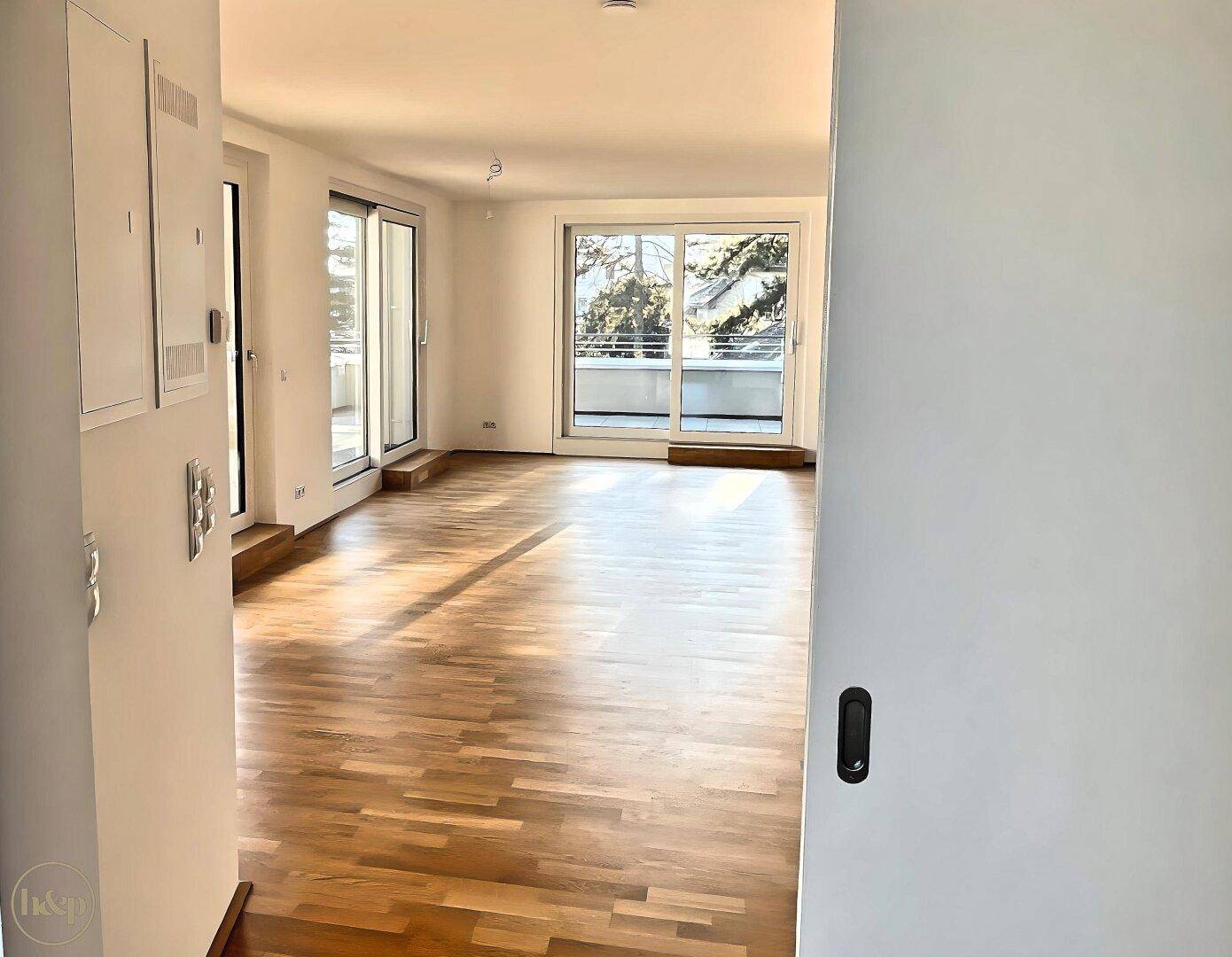 Blick vom Flur in ein helles Wohnzimmer mit Holzboden und großen Fenstern.