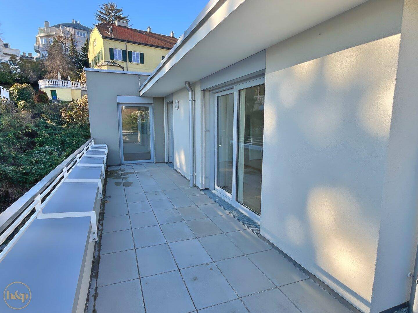 Lange, geflieste Terrasse mit weißem Geländer und Blick auf die umliegenden Häuser.