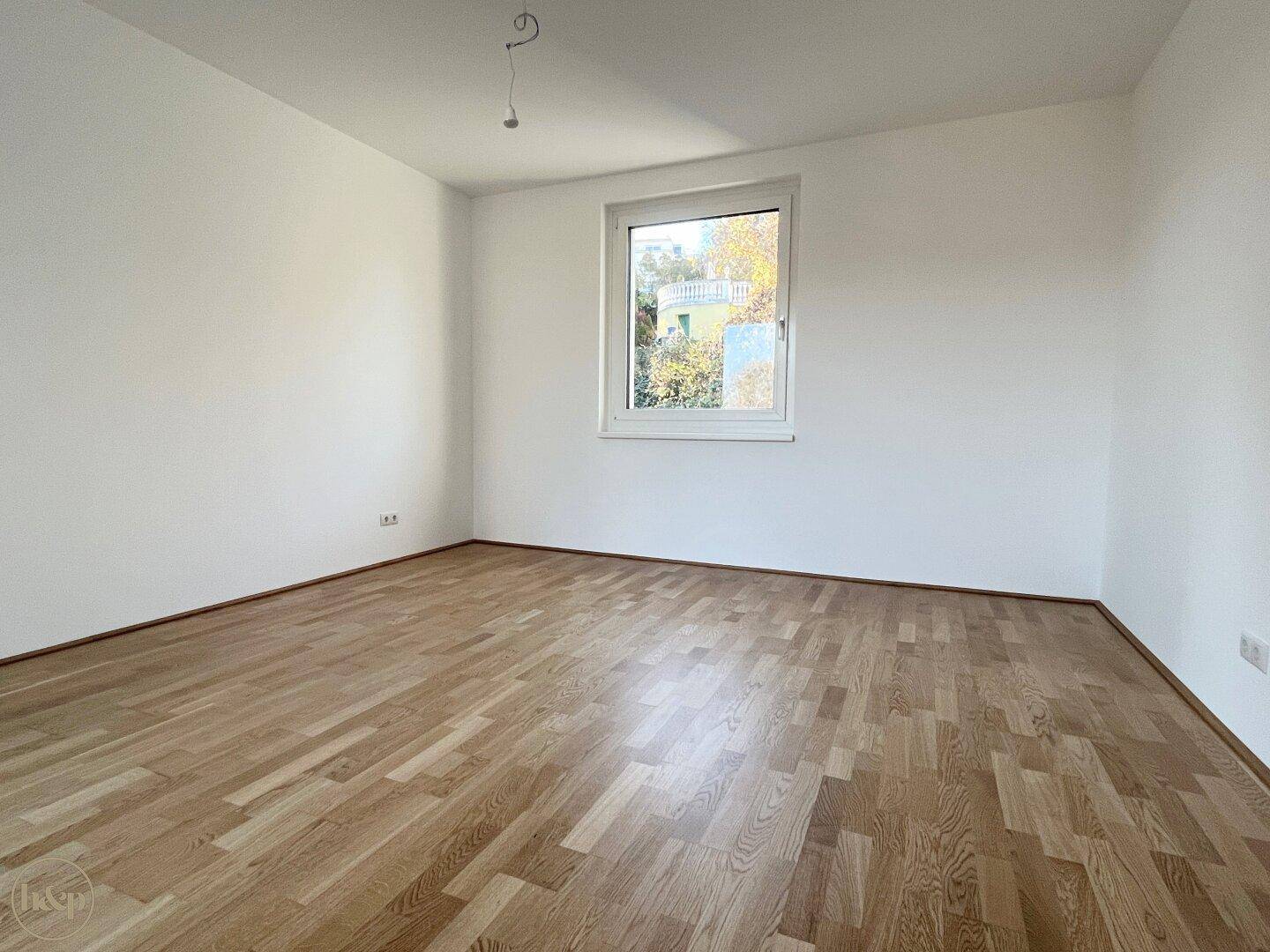 Helles, leeres Schlafzimmer mit Holzboden und einem Fenster mit Blick ins Grüne.