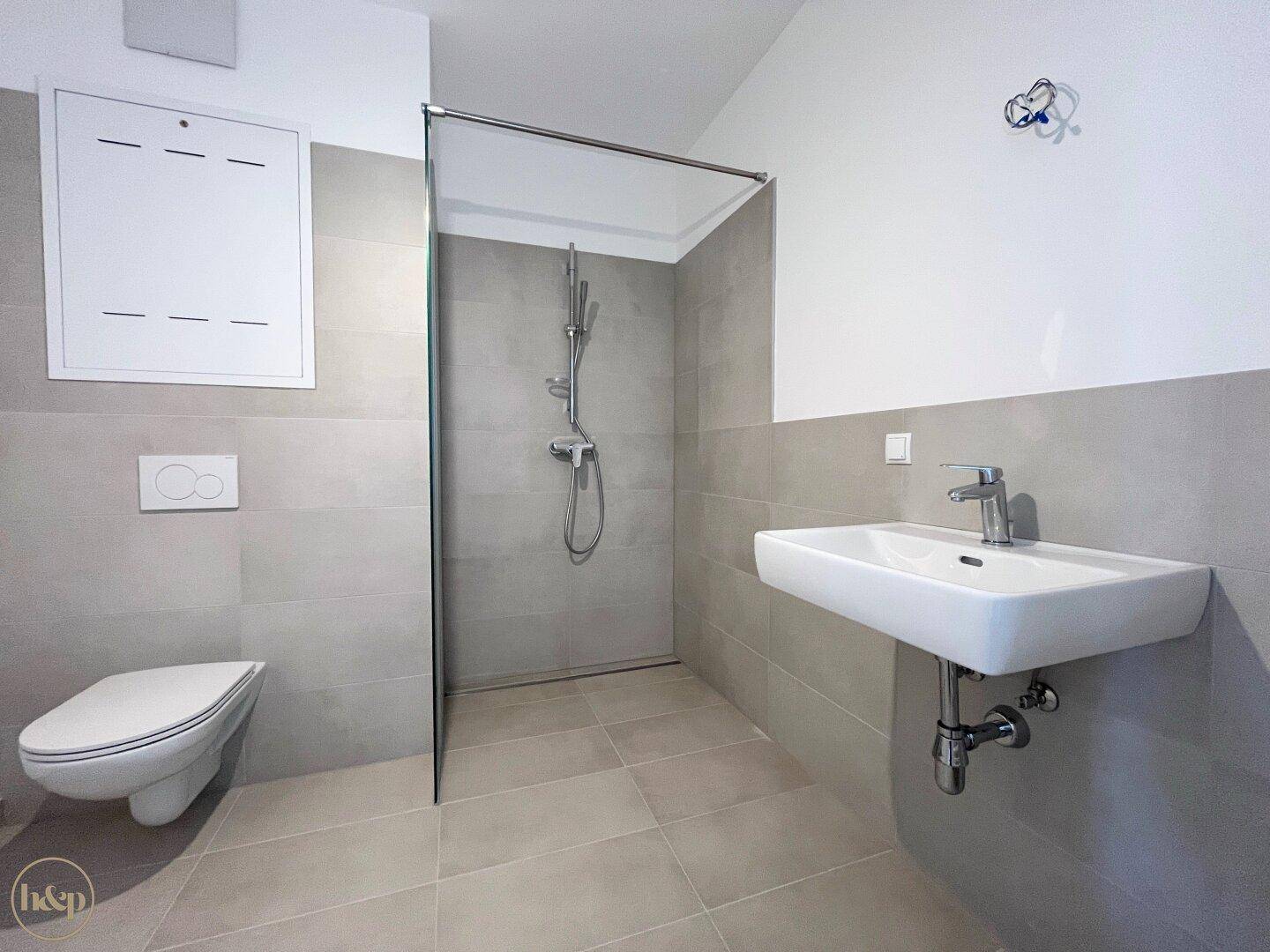 Modernes Badezimmer mit grauen Fliesen, wandhängendem WC und begehbarer Dusche.
