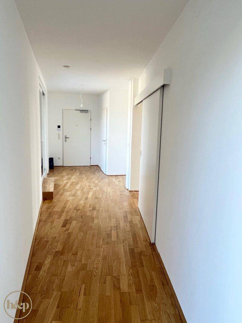 Langer, heller Flur mit Holzboden und weiß gestrichenen Wänden, der zum Eingangsbereich führt.