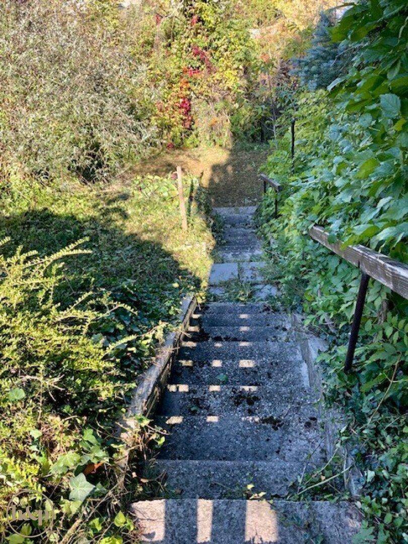 Lange, alte Steintreppe, die von üppigem Grün umgeben ist und zu einem unteren Gartenbereich führt.