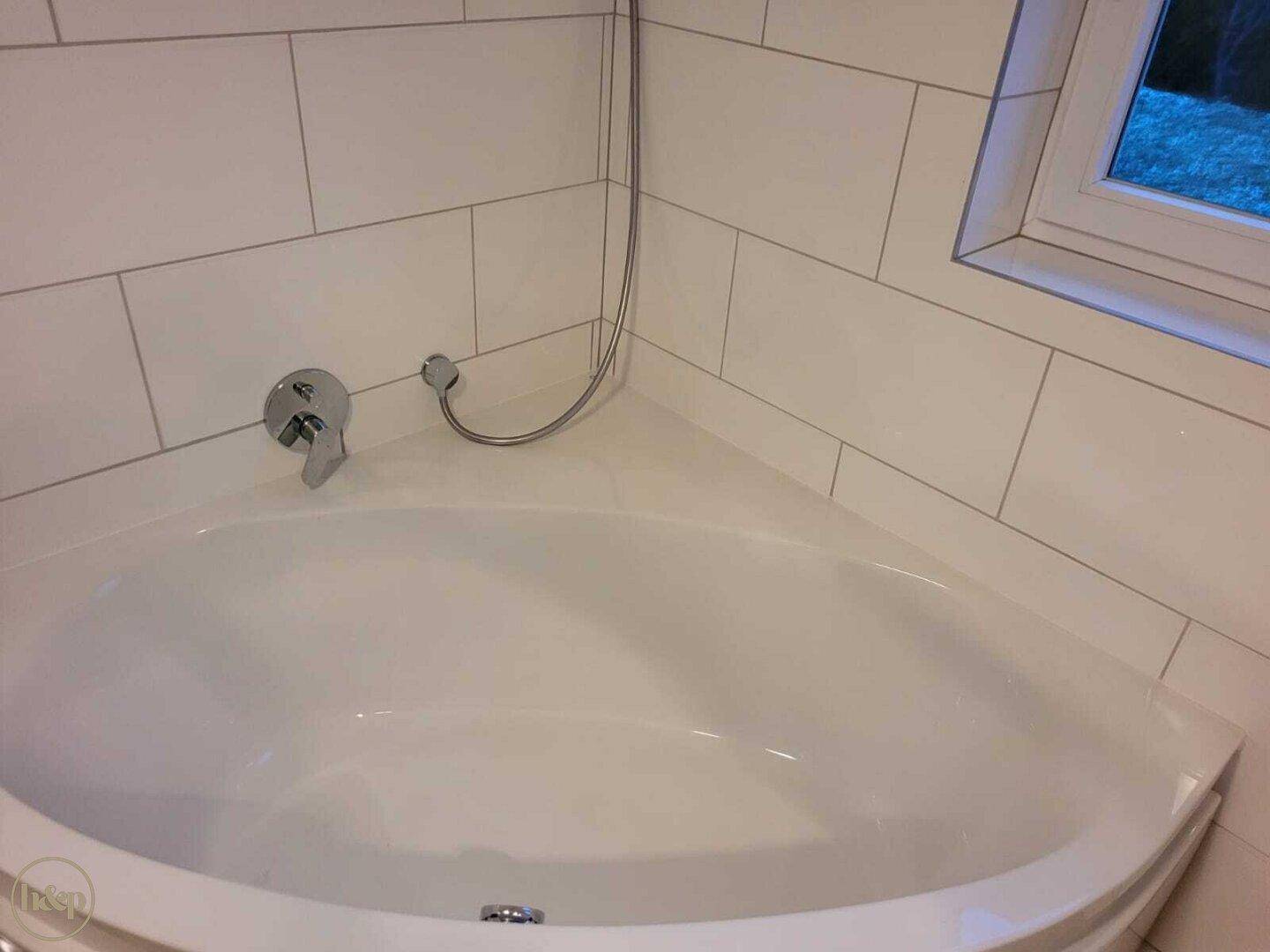 Badezimmer mit Eckbadewanne, weißen Wandfliesen und Fenster für Tageslicht.