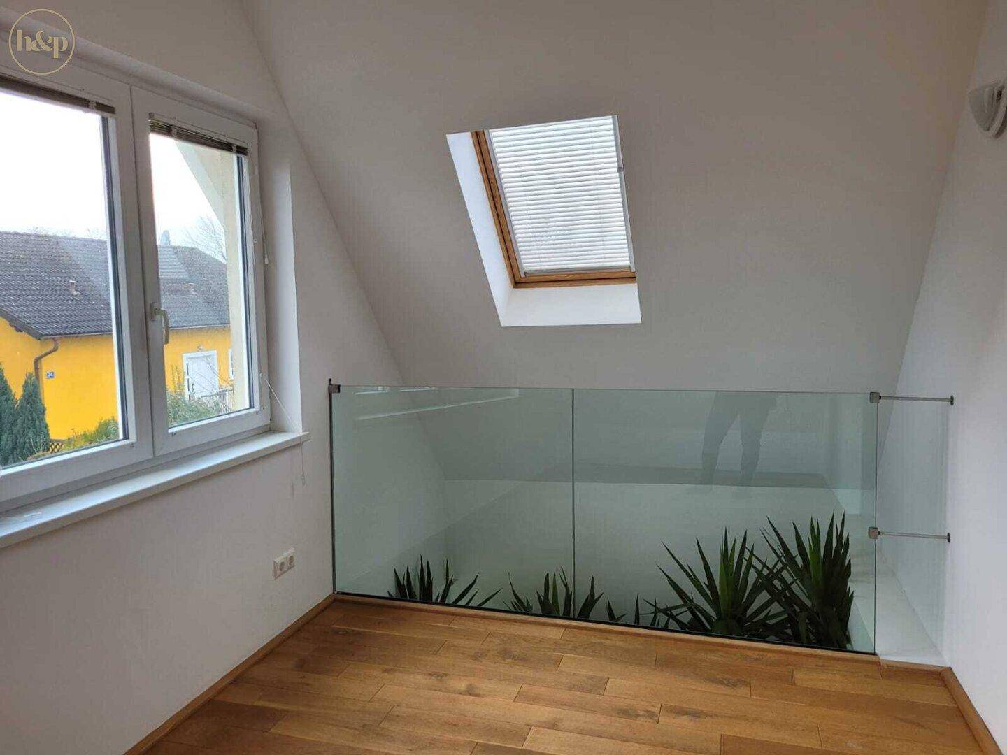 Heller Raum im Dachgeschoss mit Glasgeländer, Holzboden und Blick auf Pflanzen.