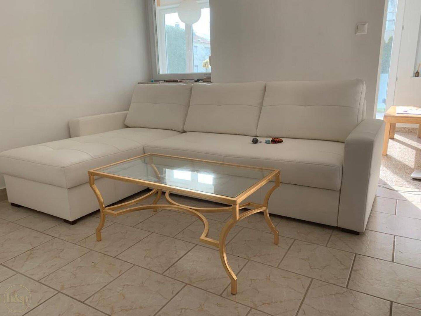 Wohnzimmer mit weißem Ecksofa, goldenem Couchtisch und Fliesenboden.