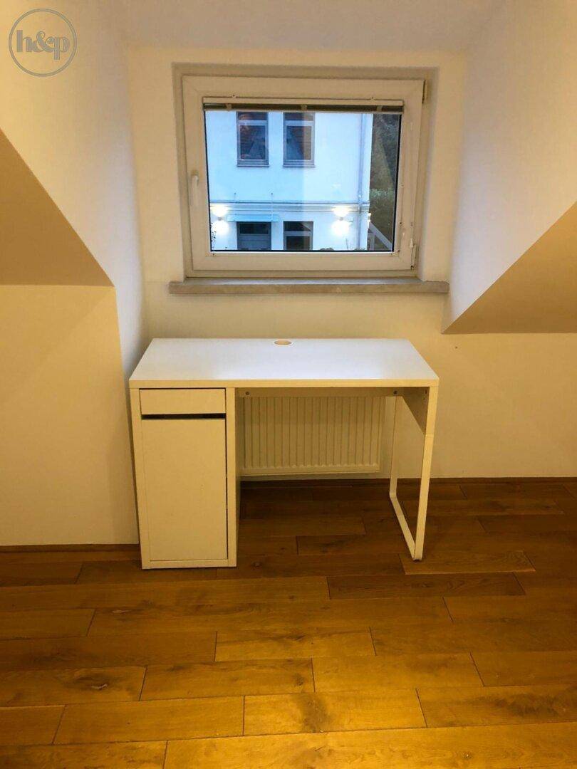 Heller Arbeitsbereich im Dachgeschoss mit weißem Schreibtisch und Fenster auf Holzboden.