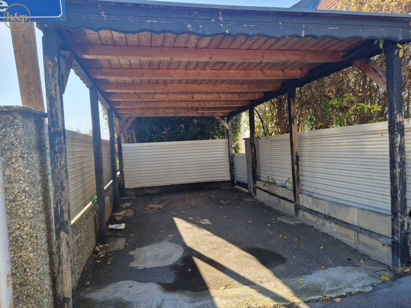 Einfacher Holz-Carport mit unbefestigtem Boden, bietet Schutz für ein Fahrzeug.
