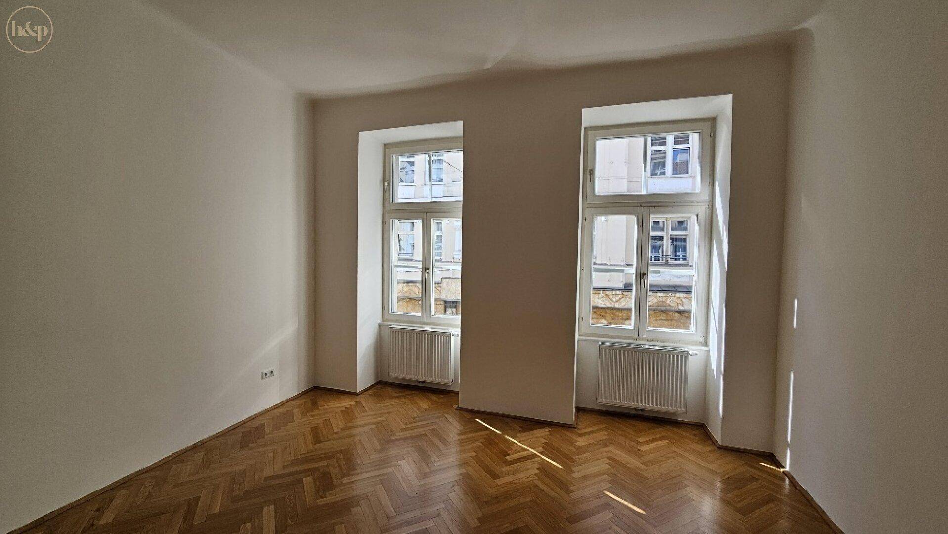 Geräumiges Zimmer mit Fischgrätparkett und zwei Fenstern, die viel Tageslicht hereinlassen.