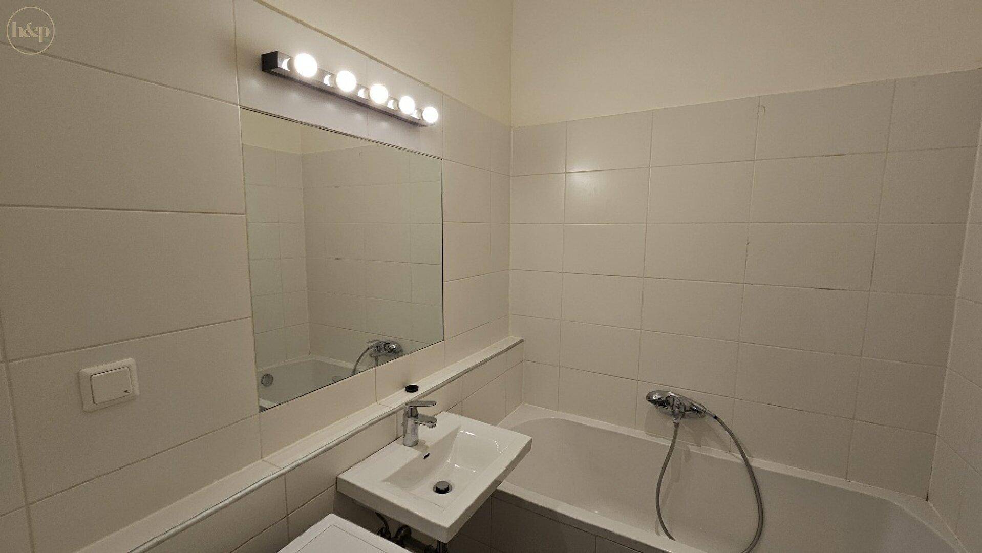 Badezimmer mit weißen Fliesen, Badewanne, Waschbecken und beleuchtetem Spiegel.