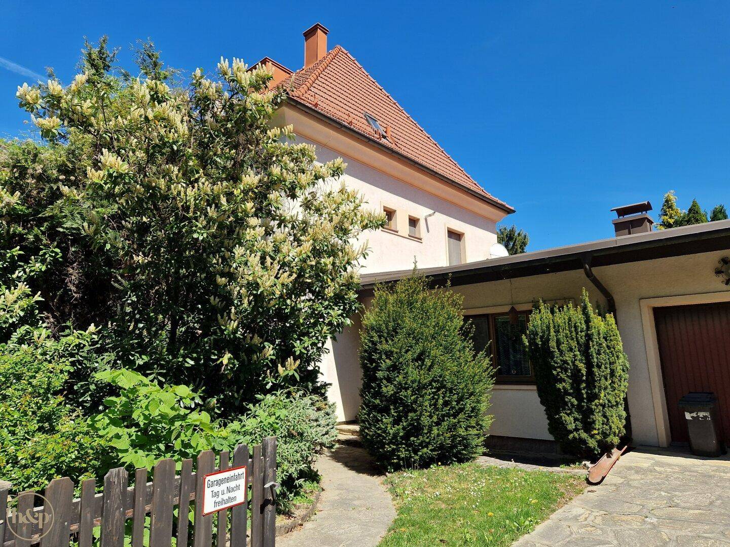 Seitenansicht des Hauses mit Garage, üppiger Bepflanzung und sonnigem Wetter.