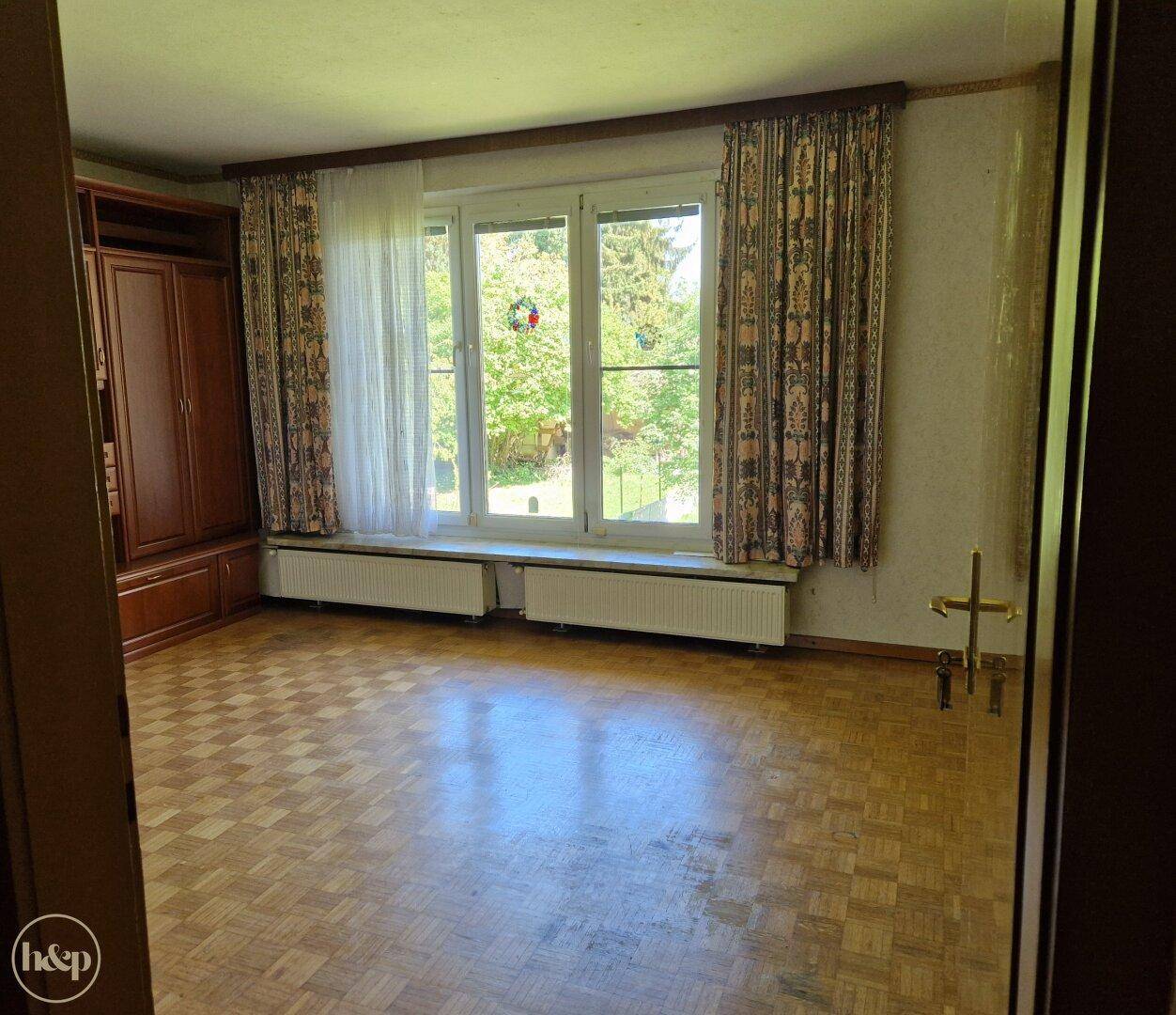 Geräumiges Zimmer mit Parkettboden, großen Fenstern und Blick ins Grüne.
