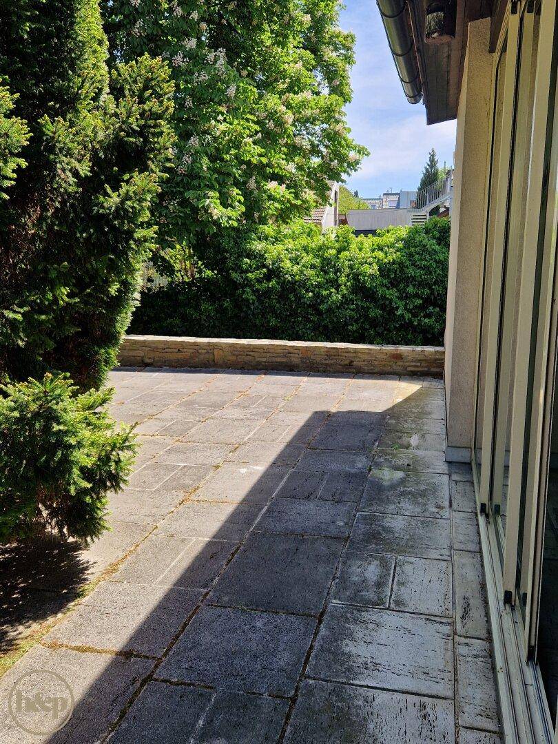 Geräumige gepflasterte Terrasse mit niedriger Steinmauer und üppiger Bepflanzung im Sonnenlicht.