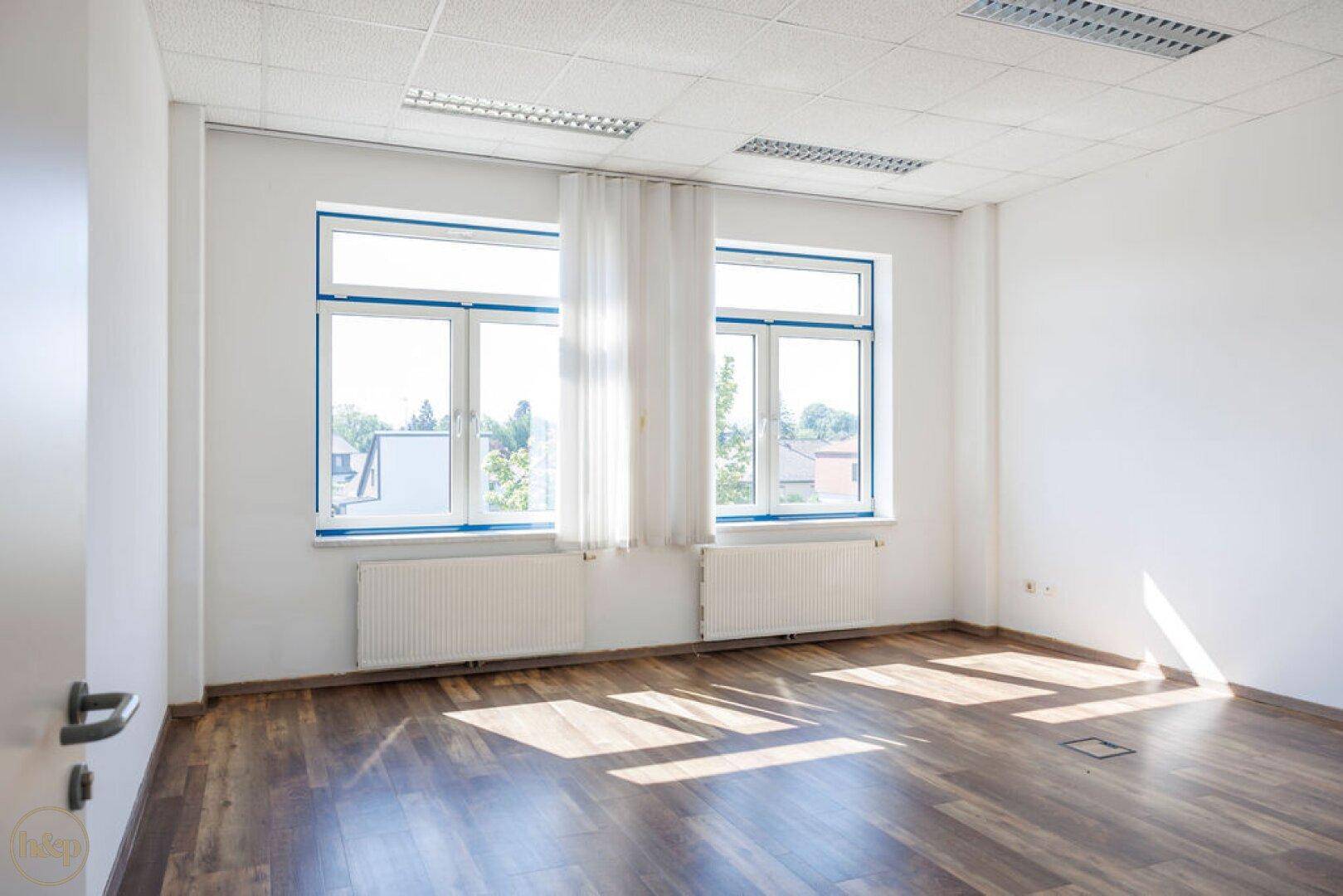 Leeres Büro mit hellen Wänden, großen Fenstern und dunklem Laminatboden.