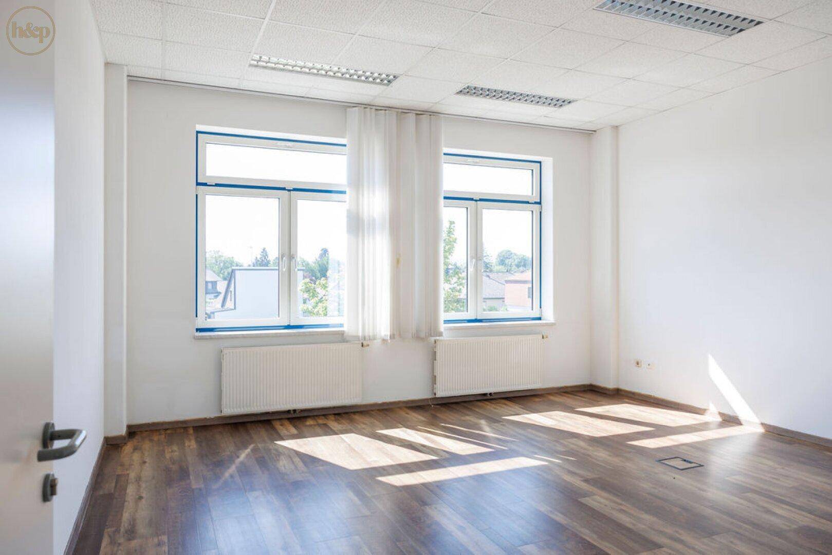 Leeres Büro mit großen Fenstern, Heizkörpern und dunklem Holzboden, bereit zur individuellen Gestaltung.
