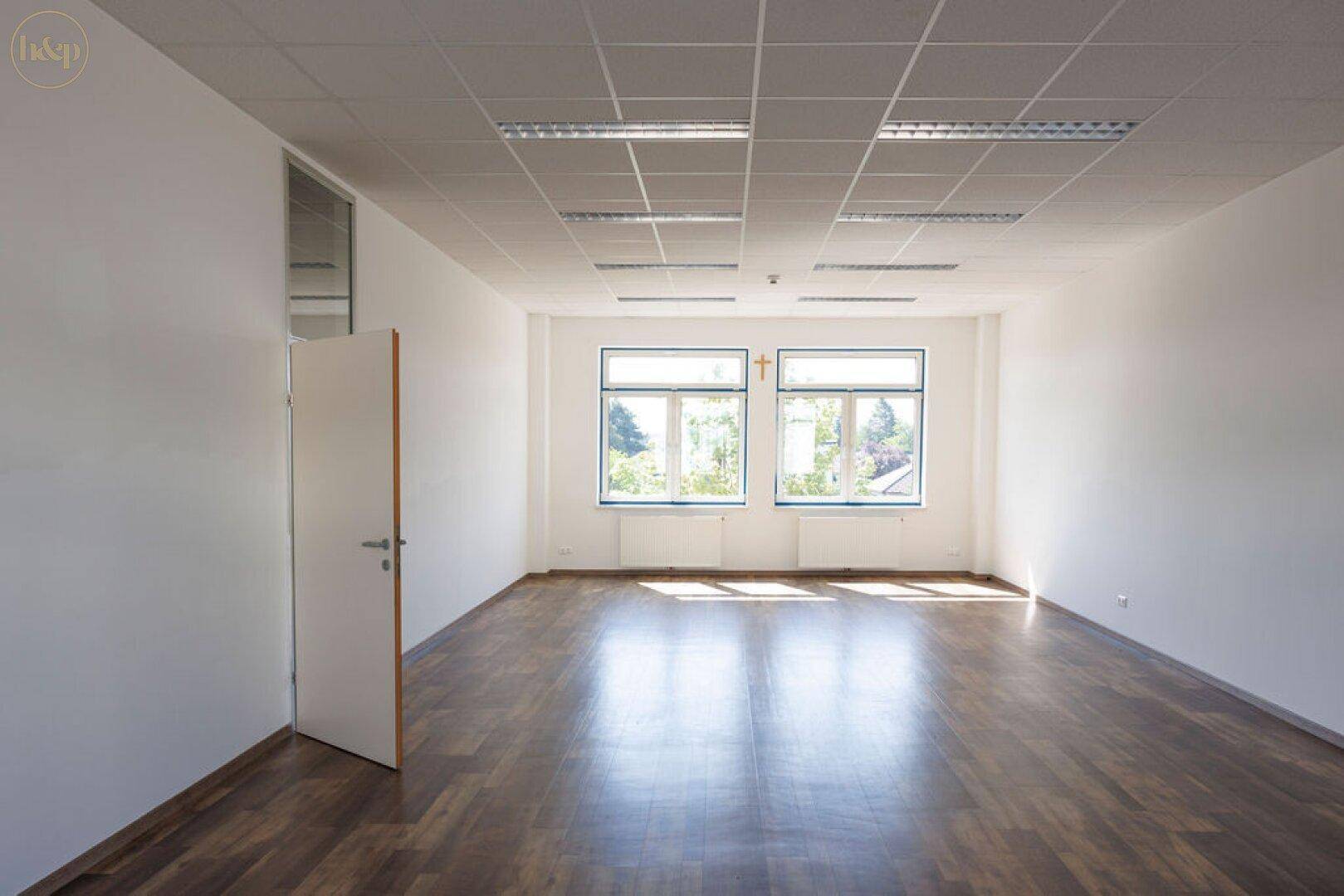 Geräumiges, leeres Büro mit großen Fenstern, Holzboden und viel Tageslicht, ideal für flexible Nutzung.