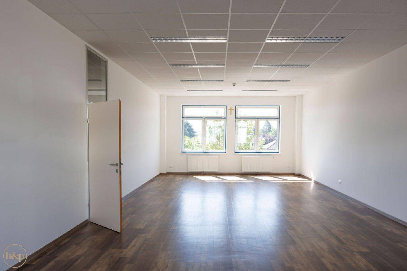 Geräumiges, leeres Büro mit hellen Wänden, großen Fenstern und dunklem Laminatboden.
