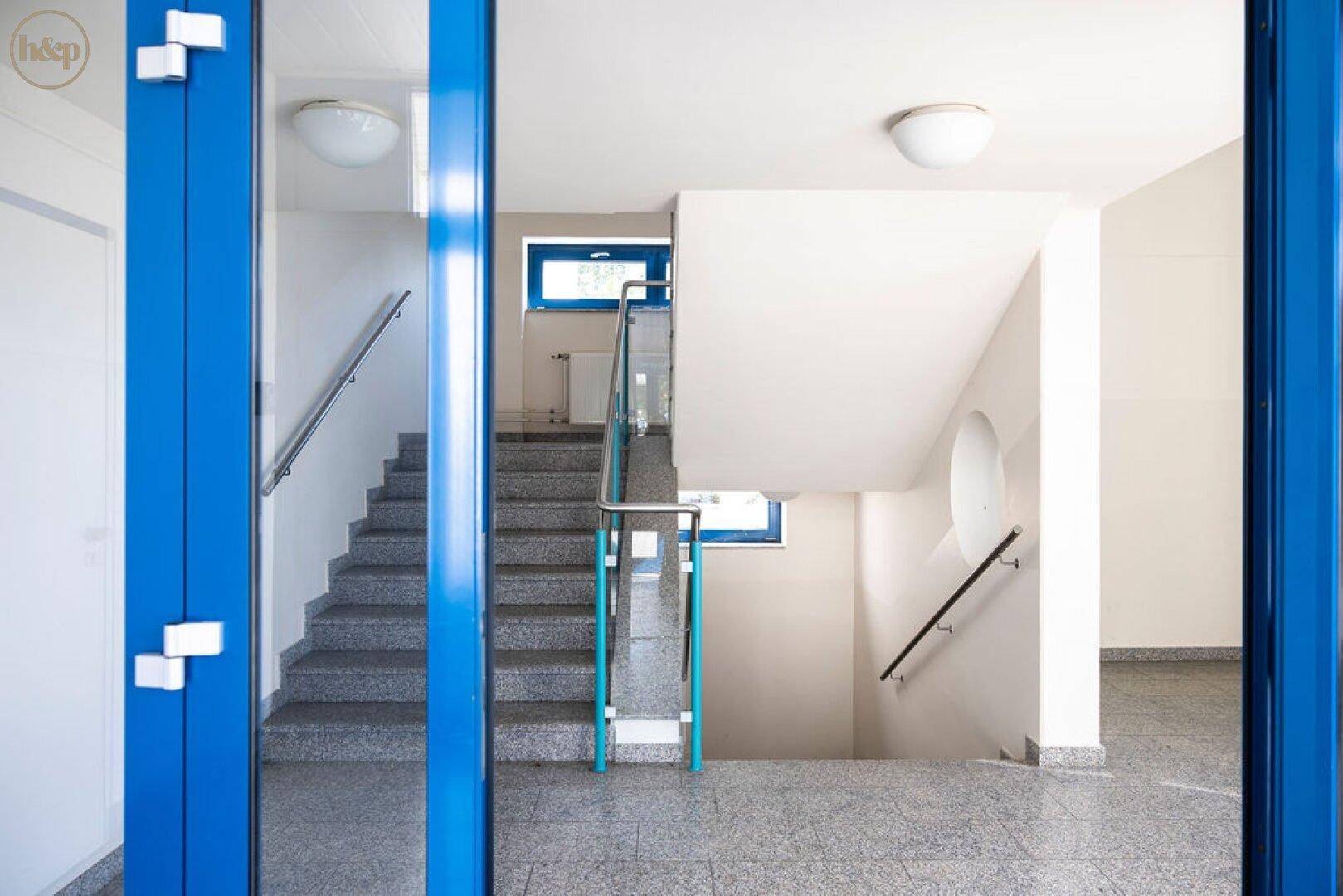 Treppenhaus mit blauen Akzenten, Glasgeländer und Terrazzoboden, hell und modern gestaltet.