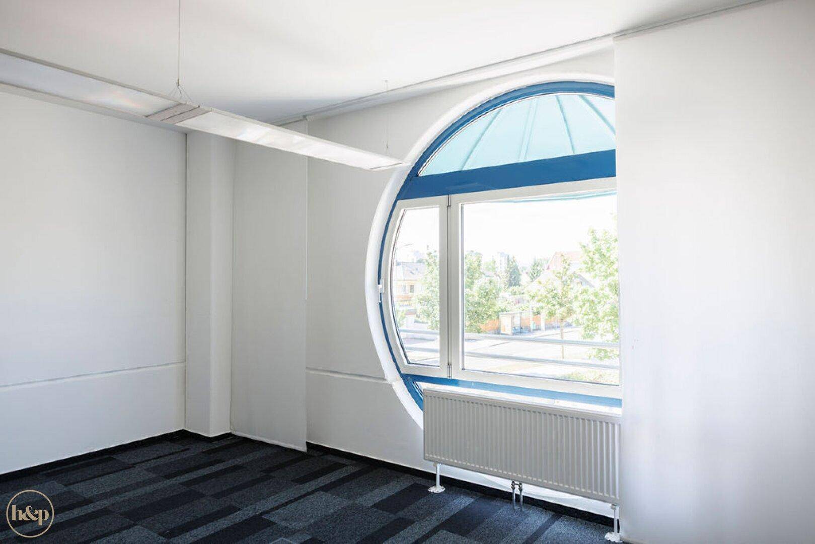 Heller, leerer Büroraum mit markantem Rundfenster und Teppichboden, bereit zur individuellen Gestaltung.