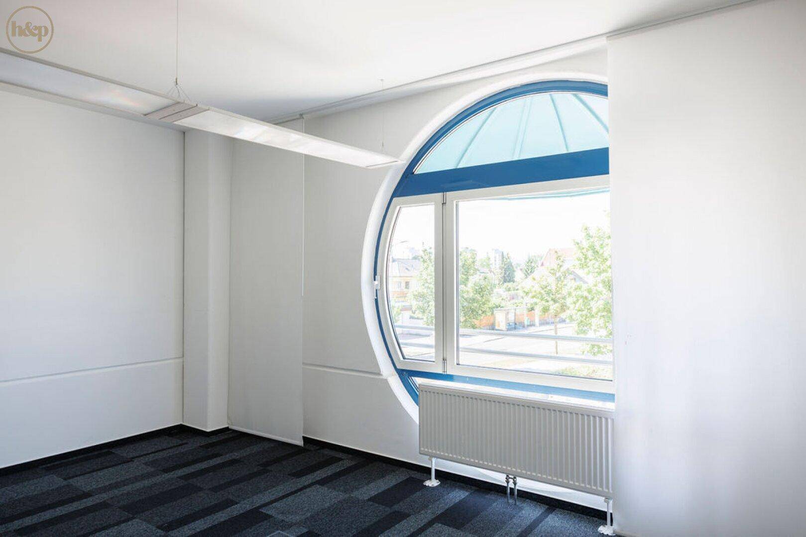 Heller Büroraum mit einzigartigem Rundfenster, Teppichboden und Heizkörper unter dem Fenster.