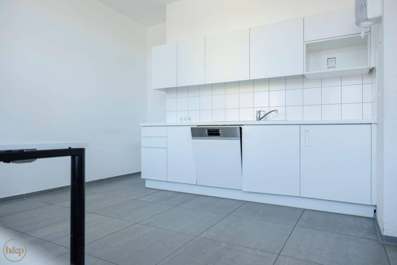 Funktionale, leere Küchenzeile mit weißen Schränken, Spülmaschine und Fliesenboden, ideal für Büronutzung.