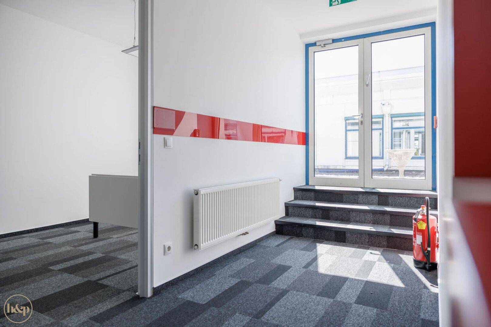 Heller Eingangsbereich mit Treppe, Glastür und Heizkörper, führt zu den Büroräumen.