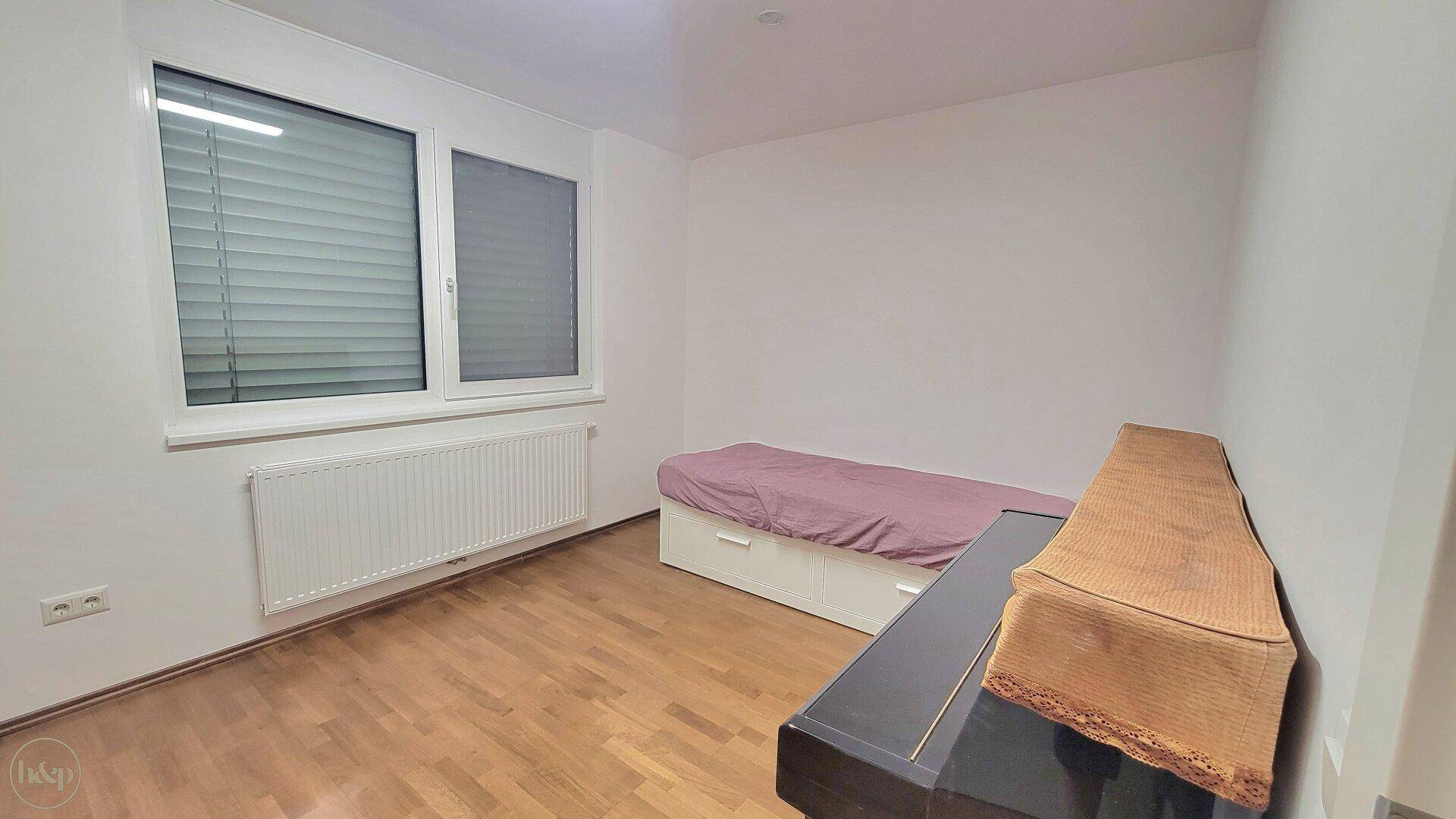 Helles Schlafzimmer mit Einzelbett, Klavier und Fenster mit Jalousien.