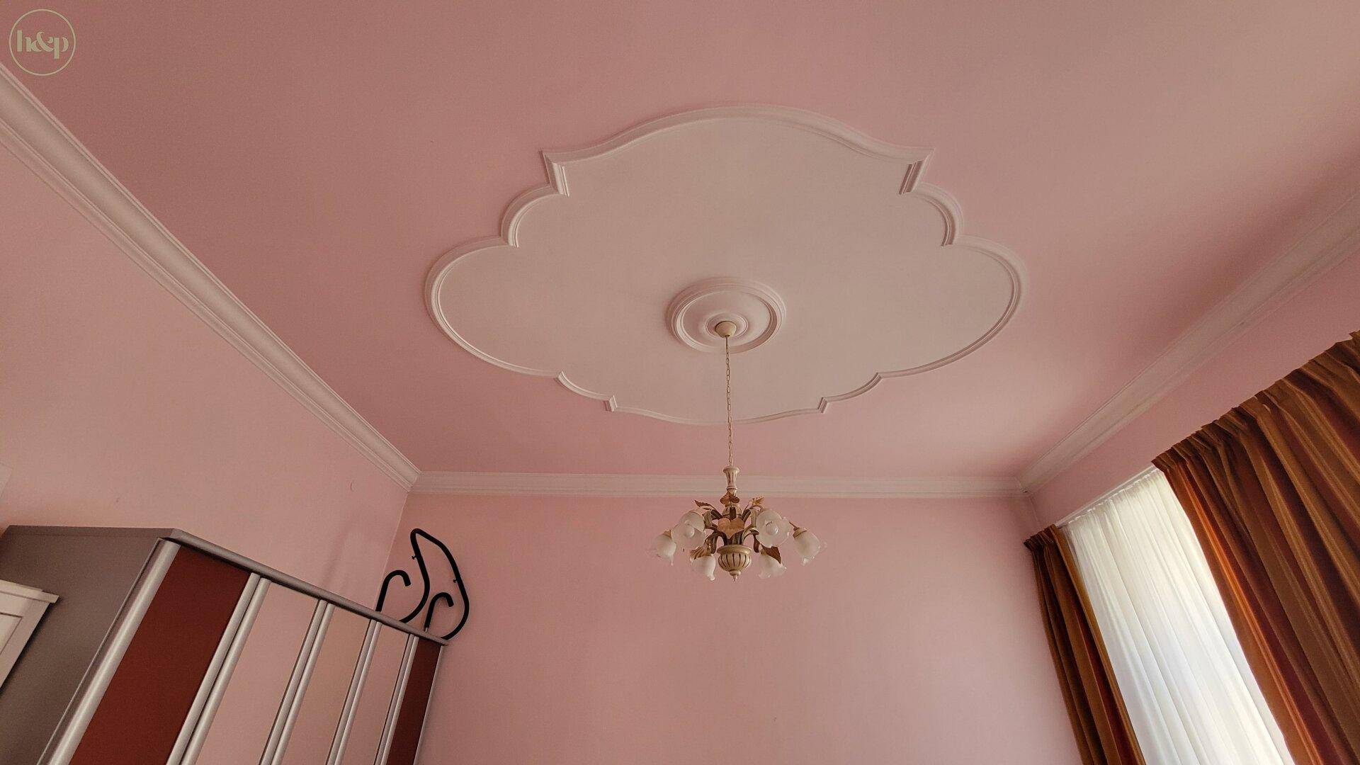 Schlafzimmer mit verzierter Stuckdecke, Kronleuchter und rosa Wänden.
