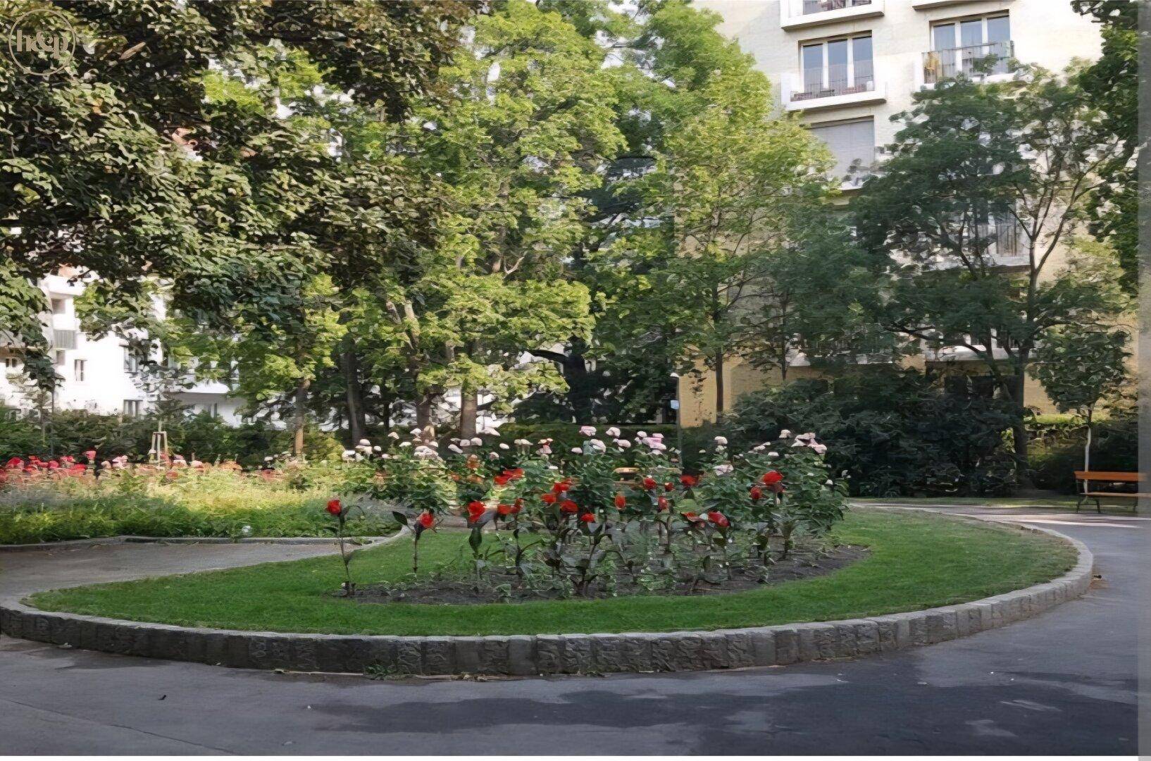 Gepflegte Grünanlage mit Rosenbeeten und hohen Bäumen vor einem Wohngebäude in städtischer Umgebung.