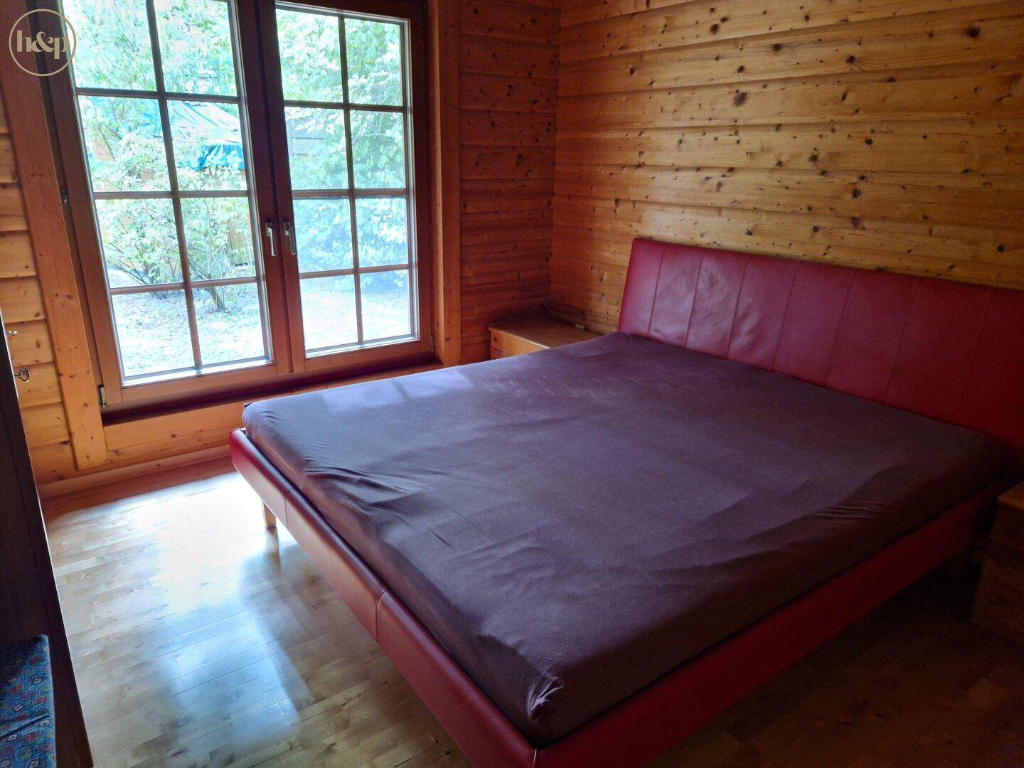 Schlafzimmer mit rotem Bettrahmen, Holzwänden und Blick ins Grüne.