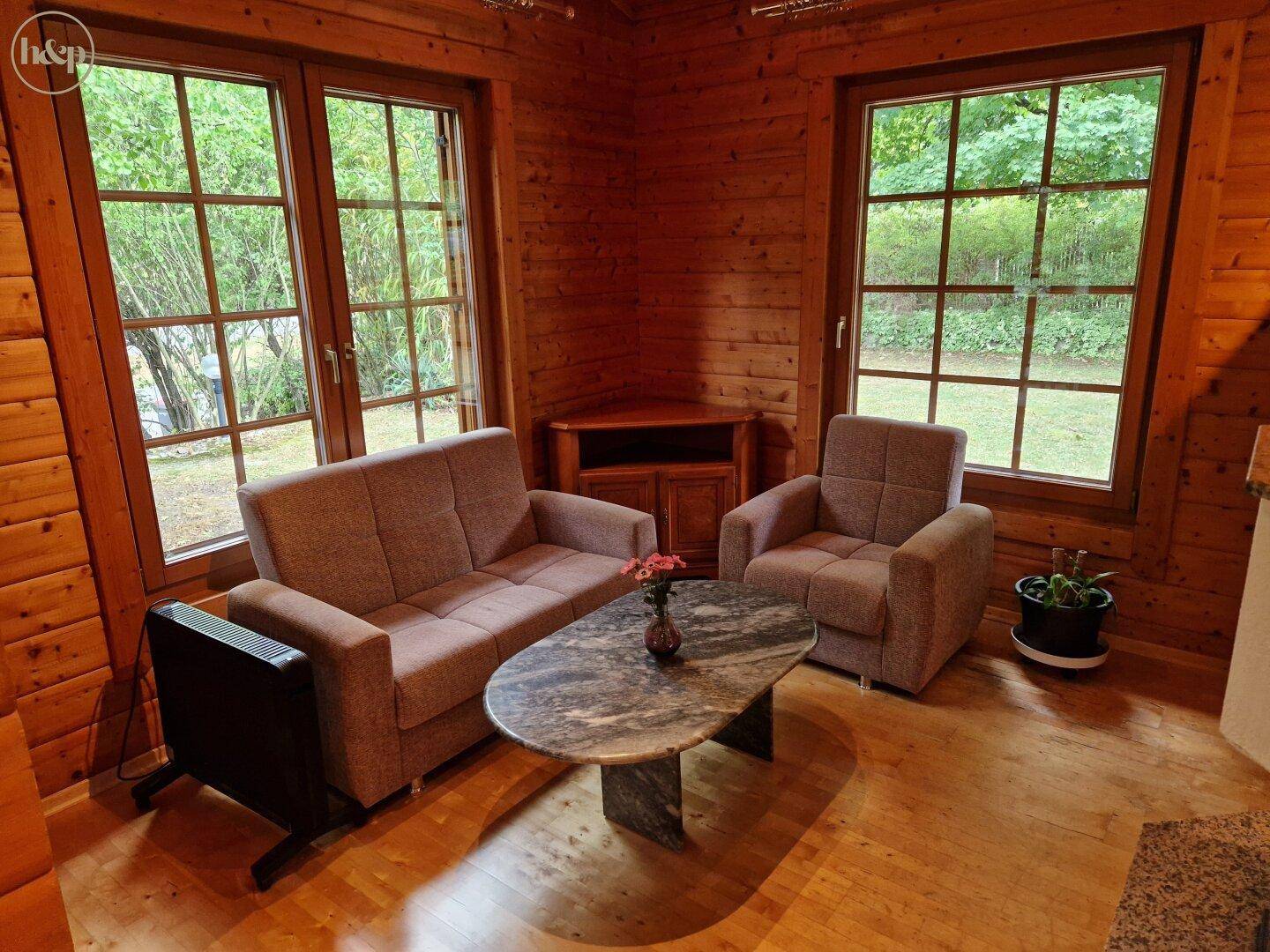 Gemütliches Wohnzimmer mit Sofa, Sessel und Holzwänden, umgeben von Natur.