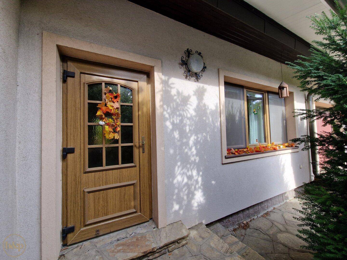 Eingangsbereich des Hauses mit einer Holztür und einem kleinen Fenster, umgeben von Putzfassade.