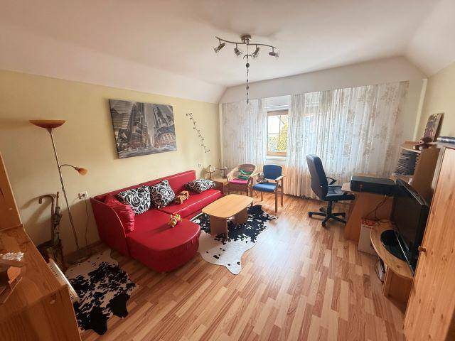Helles Zimmer mit rotem Sofa, Schreibtisch und Laminatboden unter einem Dachschrägenfenster.