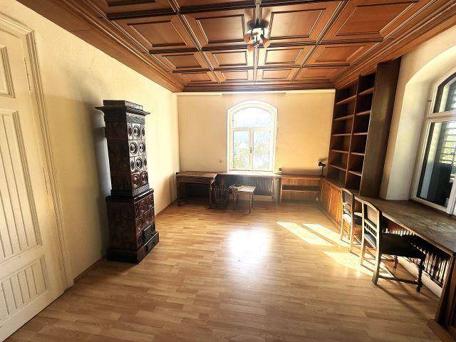Helles Zimmer mit Holzboden, Kassettendecke und einem traditionellen Kachelofen, ideal als Wohn- oder Arbeitszimmer.